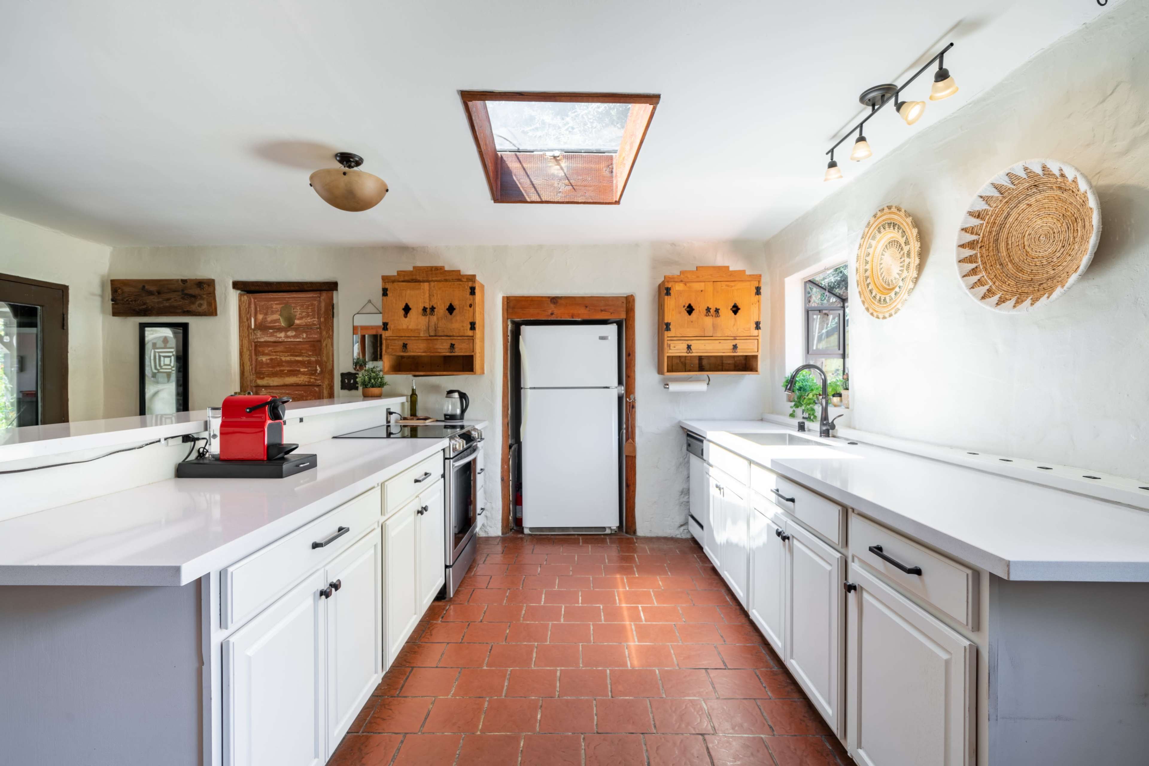 The kitchen features white cabinetry, a central skylight, and terracotta tile flooring with appliances arranged along the walls.