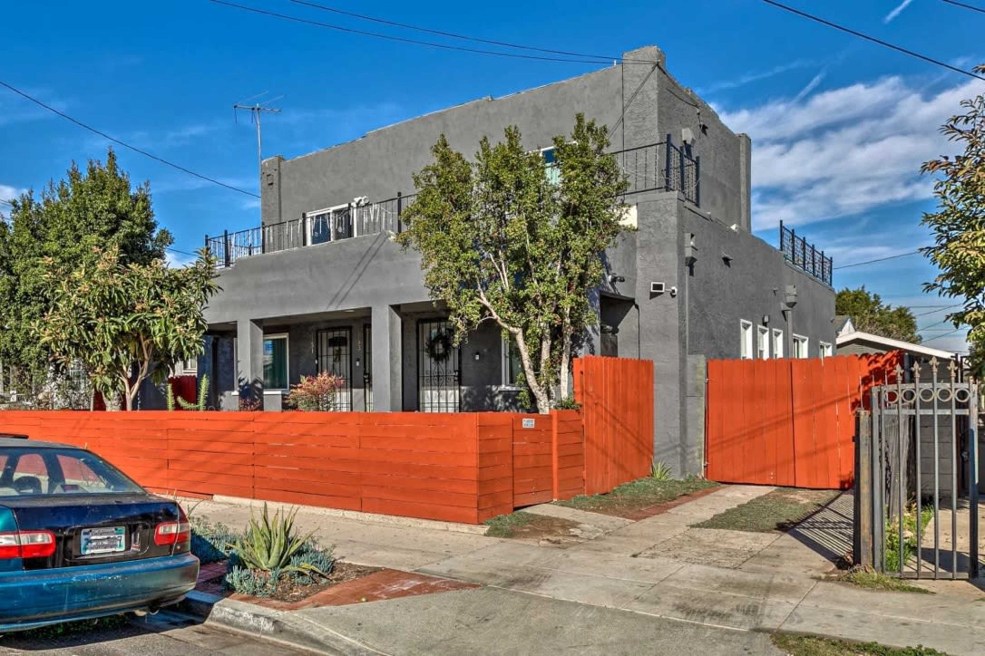 The image shows a two-story gray house with a balcony, surrounded by a bright red wooden fence and several trees, located on a neighborhood street.