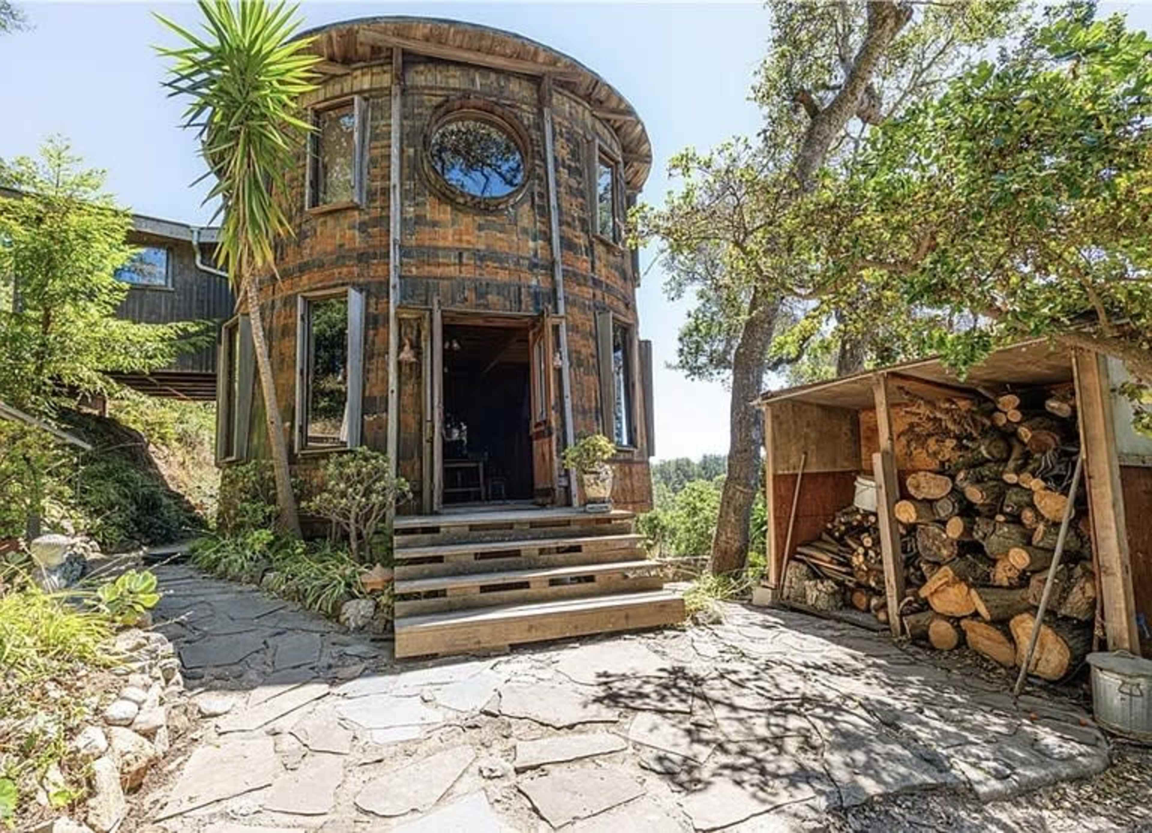 A round, wooden structure with large windows and a circular entrance is surrounded by greenery and stone pathways, with a log storage next to it.