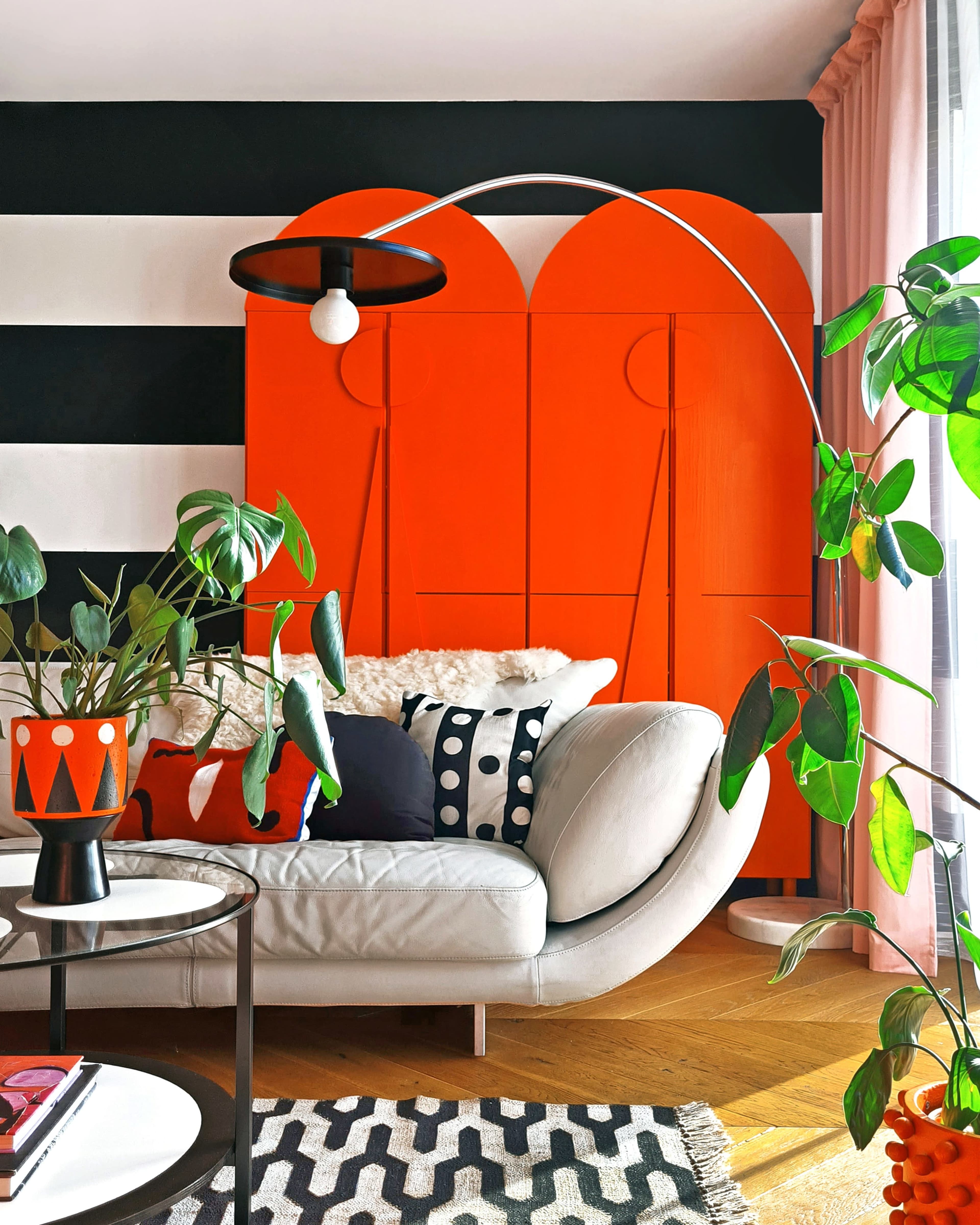 The scene features a modern living room with a curved white sofa, an orange cabinet, a coffee table, and various indoor plants against a black and white striped wall.