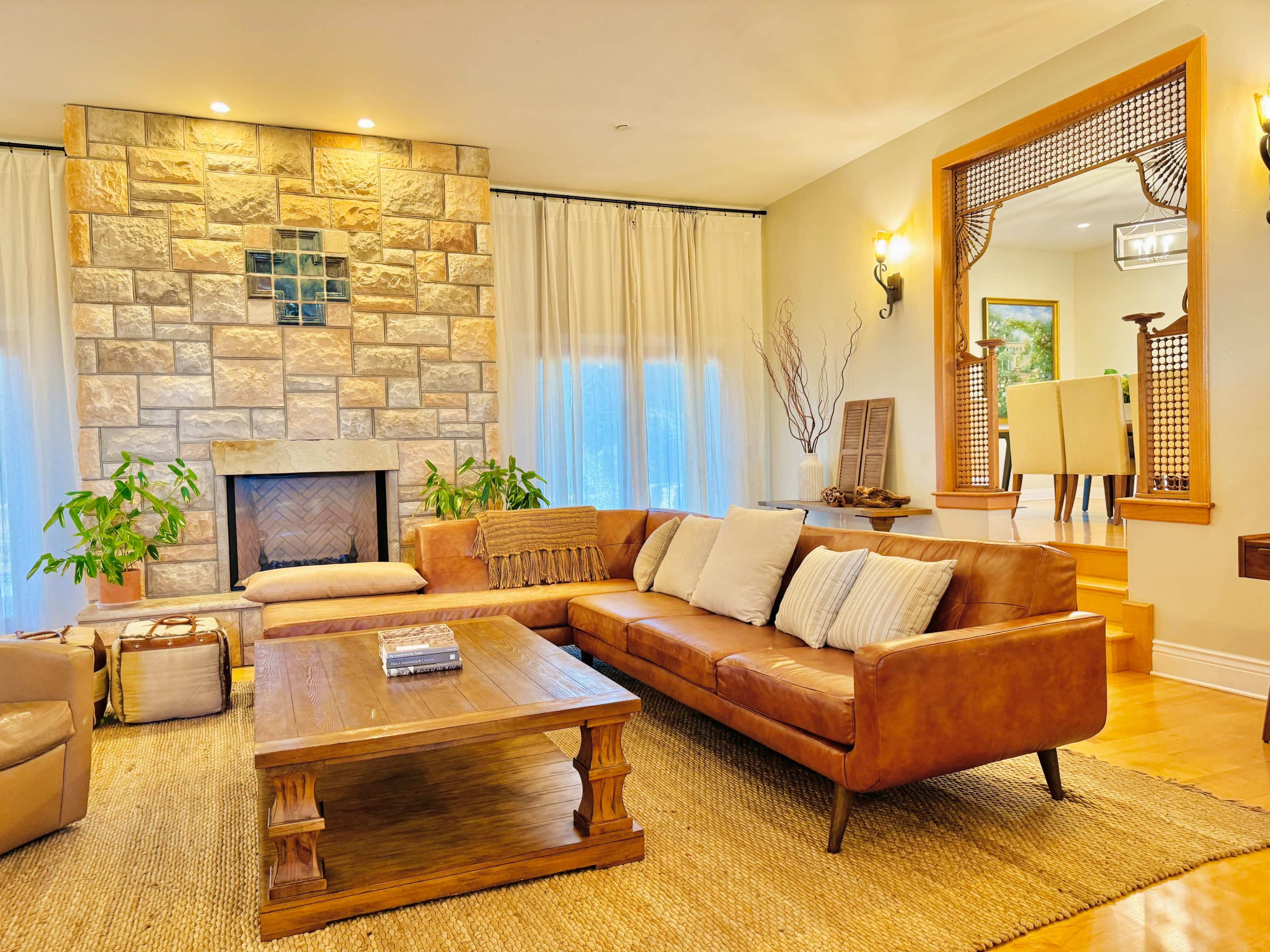 The image shows a cozy living room featuring a leather sofa, a stone fireplace, and a wooden coffee table, with large windows covered by sheer curtains.