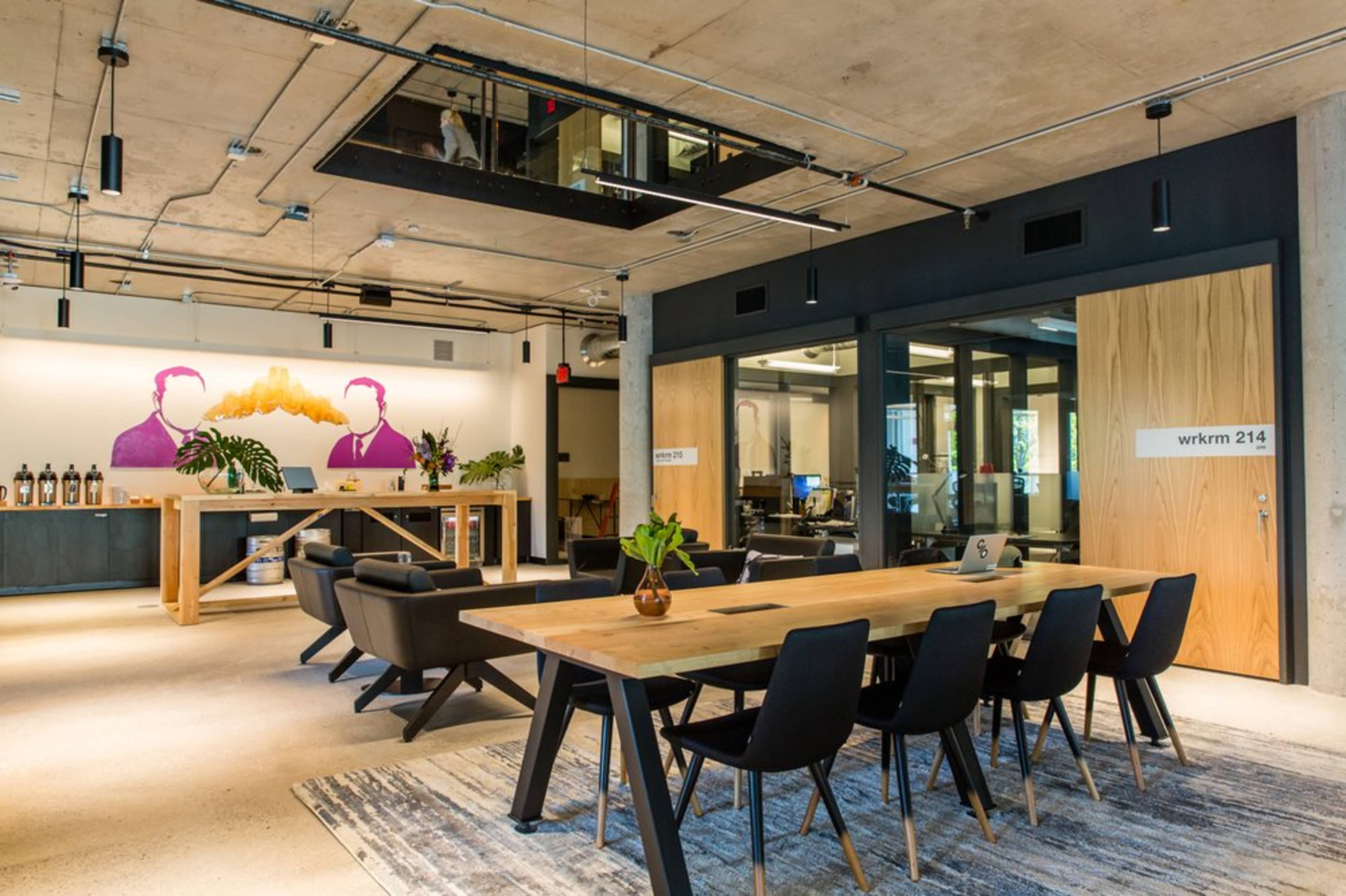 A modern office space with a large wooden table, black chairs, and a mural of two figures on the wall, alongside glass office partitions and greenery.