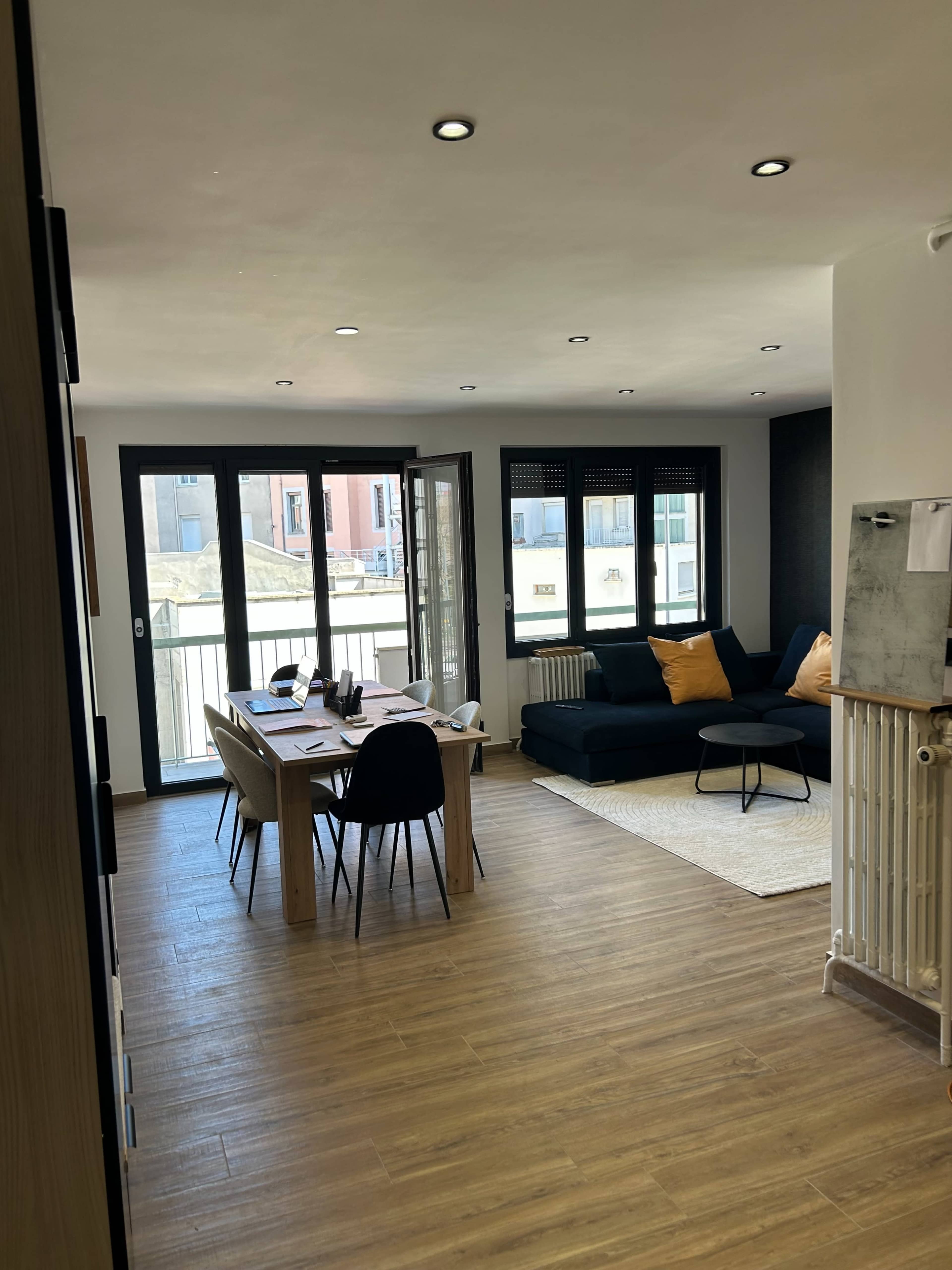 The image shows a modern living space with a dining area featuring a wooden table and black chairs, a dark sofa with orange cushions, and large windows allowing natural light to enter.