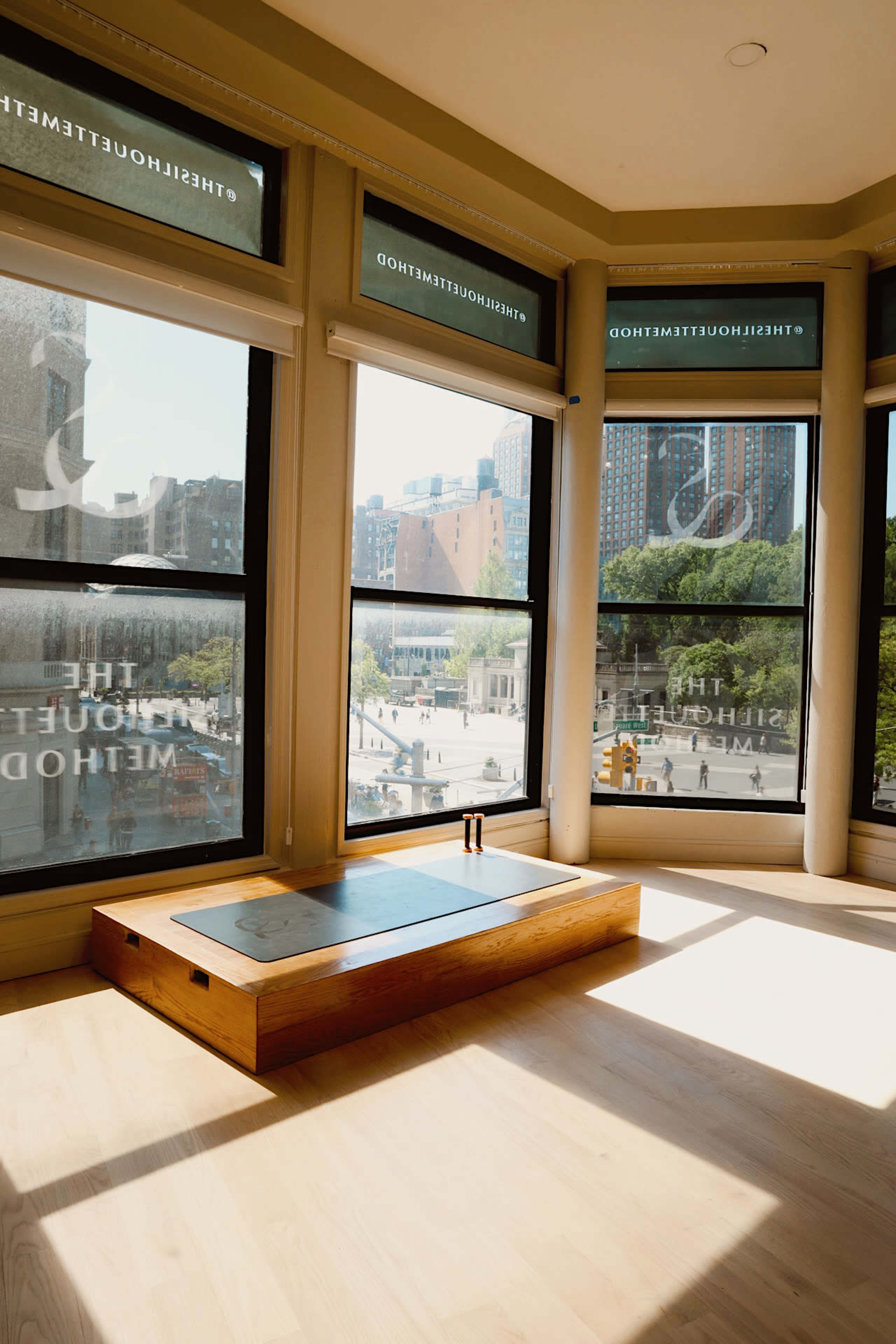 A sunlit room features a wooden platform near large windows, showcasing an urban view and decorated with minimalist furnishings.