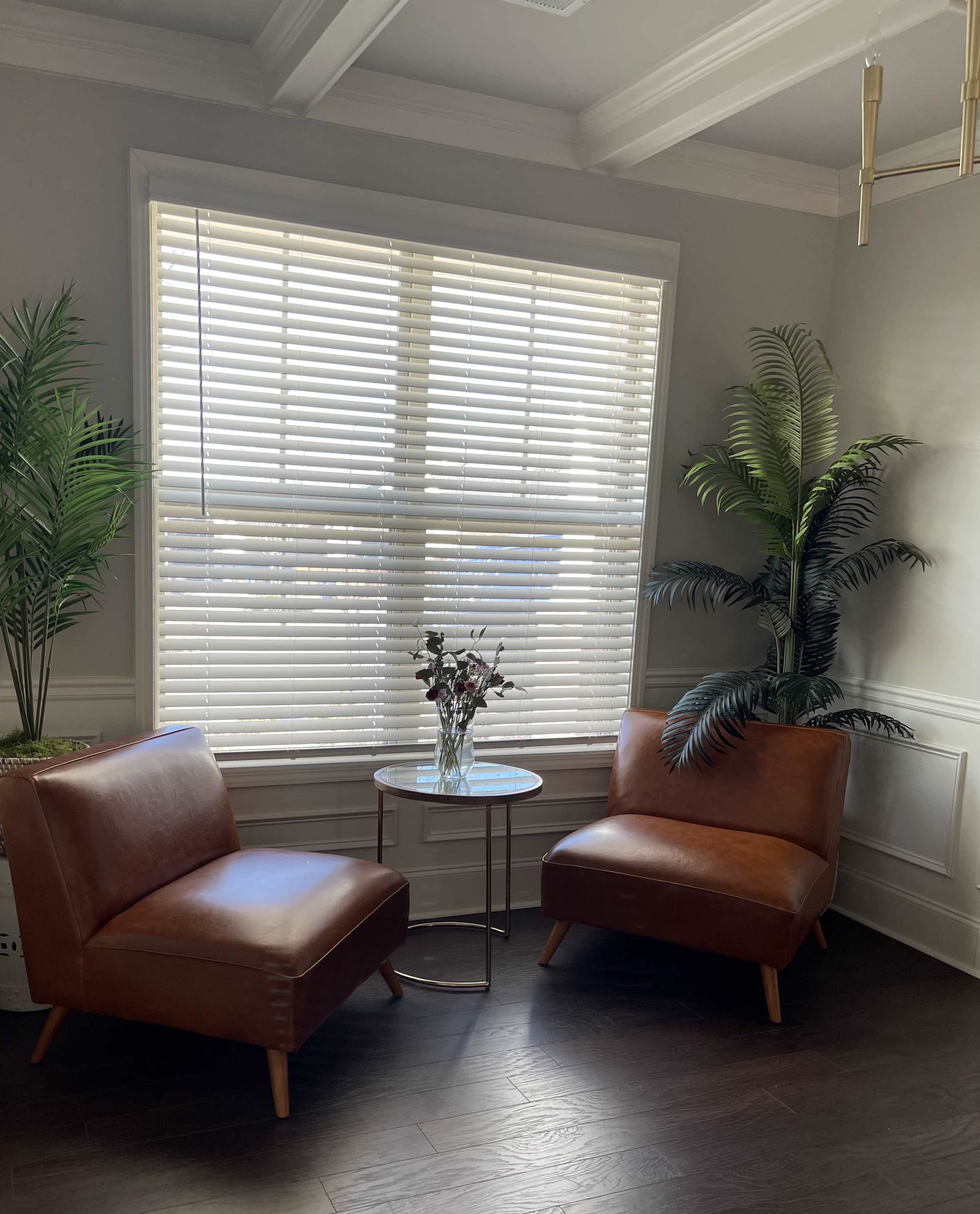 A cozy corner features two brown leather chairs and a small round table, surrounded by tall plants under a window with blinds.