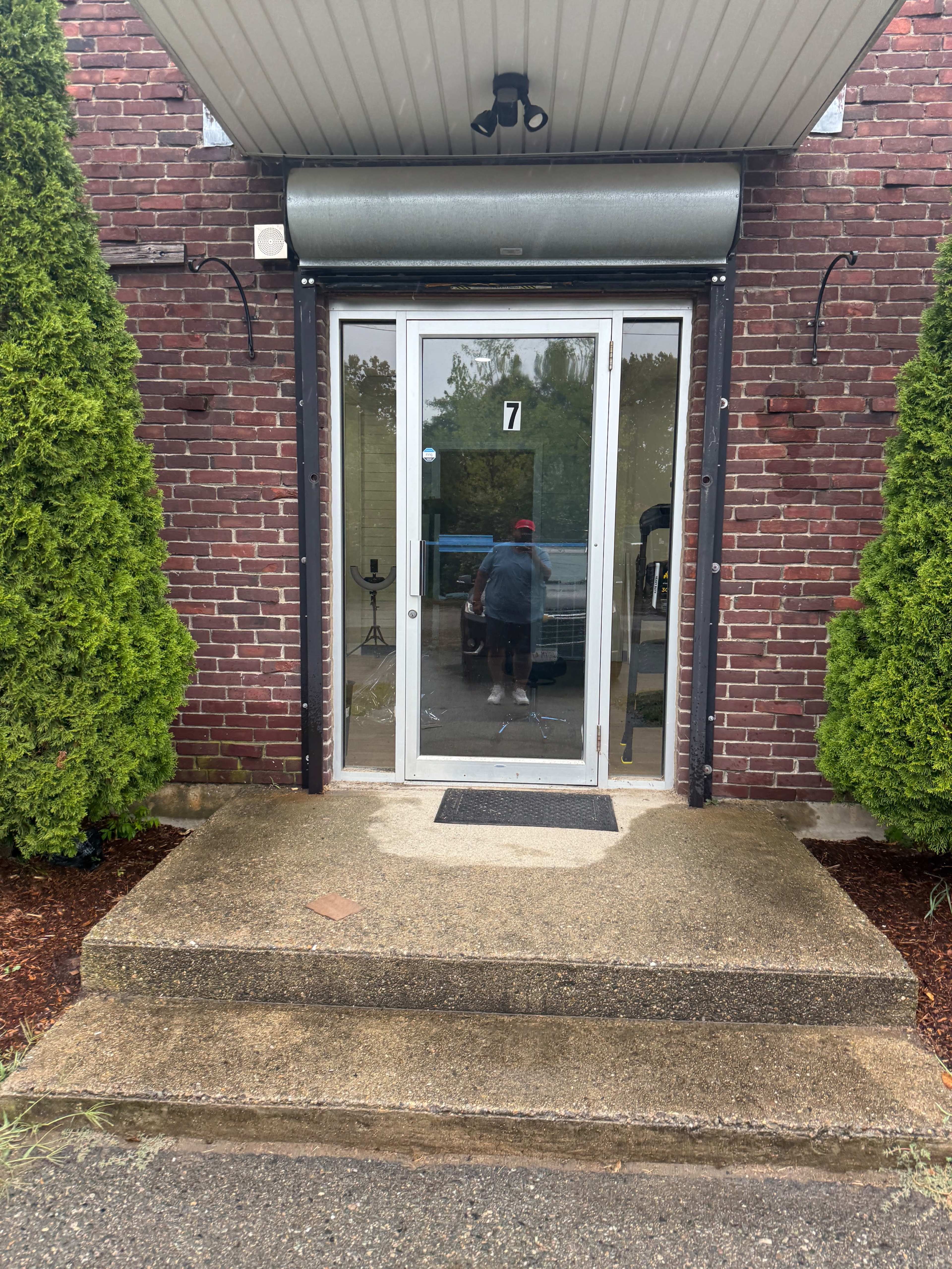 The image shows a brick building entrance with a glass door, flanked by two green bushes, and a small set of concrete steps leading up to it.