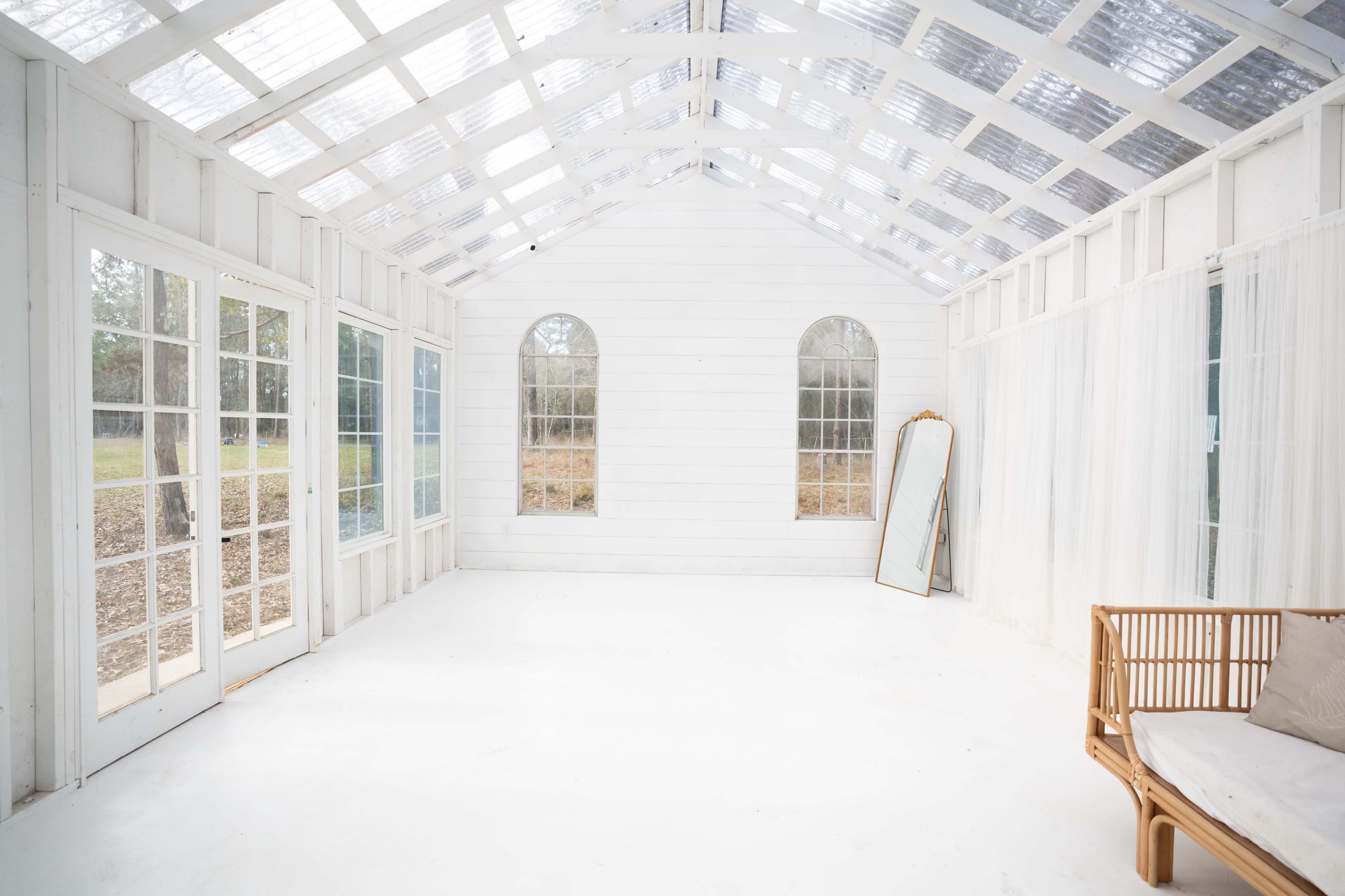 A bright, empty sunroom features white walls, large windows, and a rattan chair with a mirror leaning against the wall.