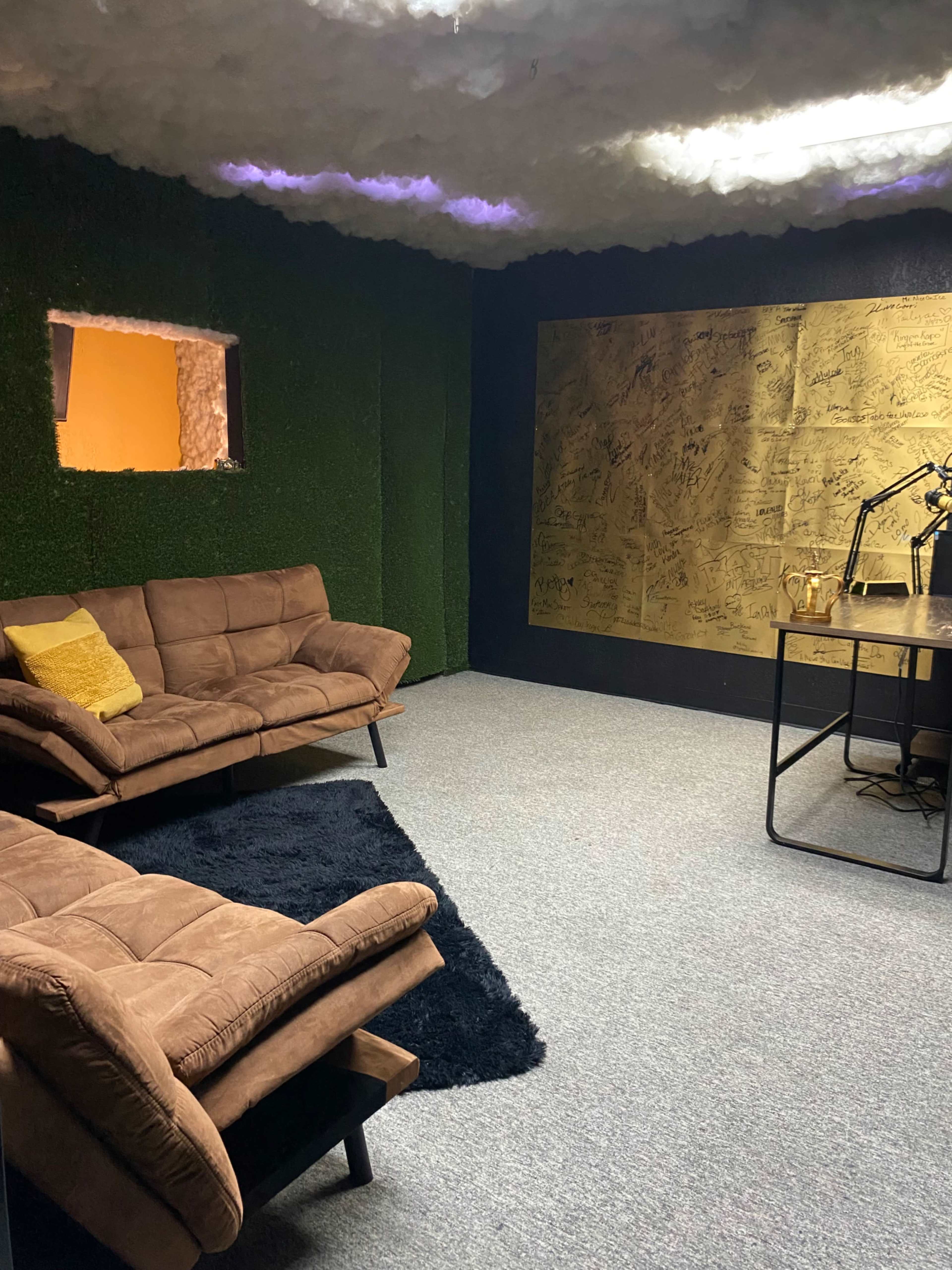 The image shows a small, modern room with dark green and brown decor, featuring two brown couches, a black rug, and a wall covered with golden notes or drawings.