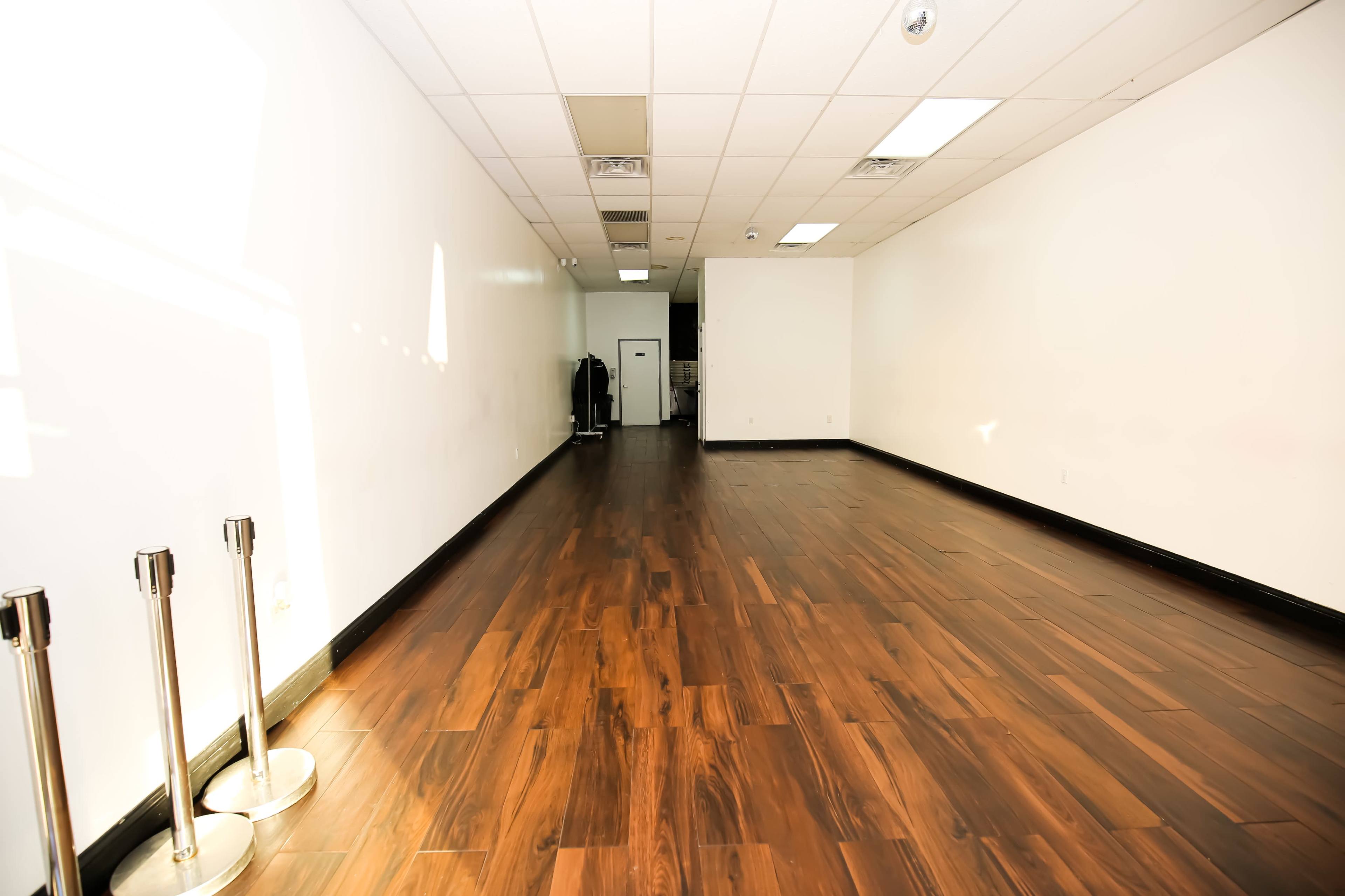 The image shows a spacious, empty room with wooden flooring and white walls, featuring a small doorway at the far end.