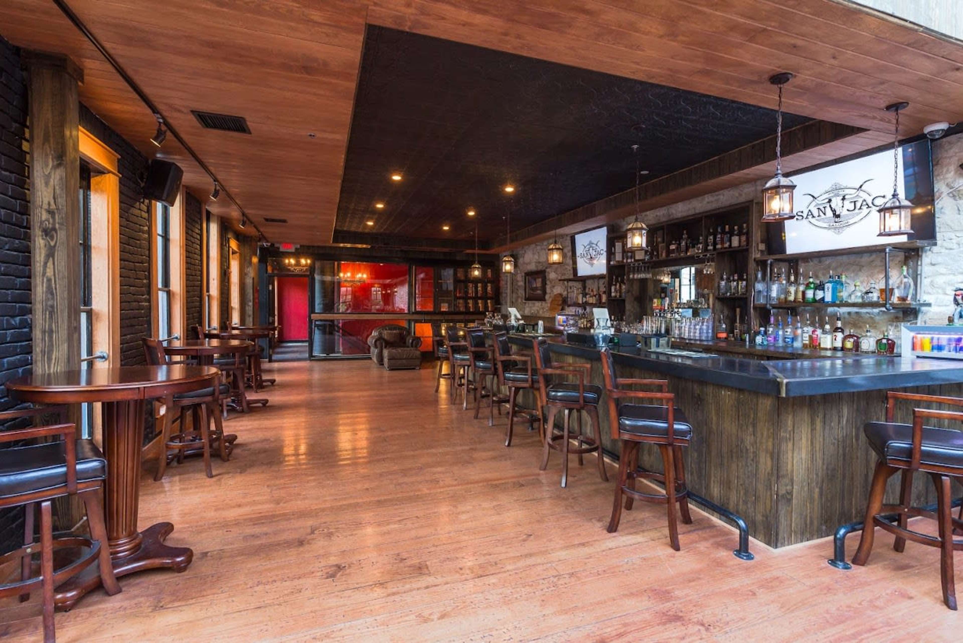 A bar interior with wooden floors, a large bar counter, and seating arrangements of tables and high chairs.