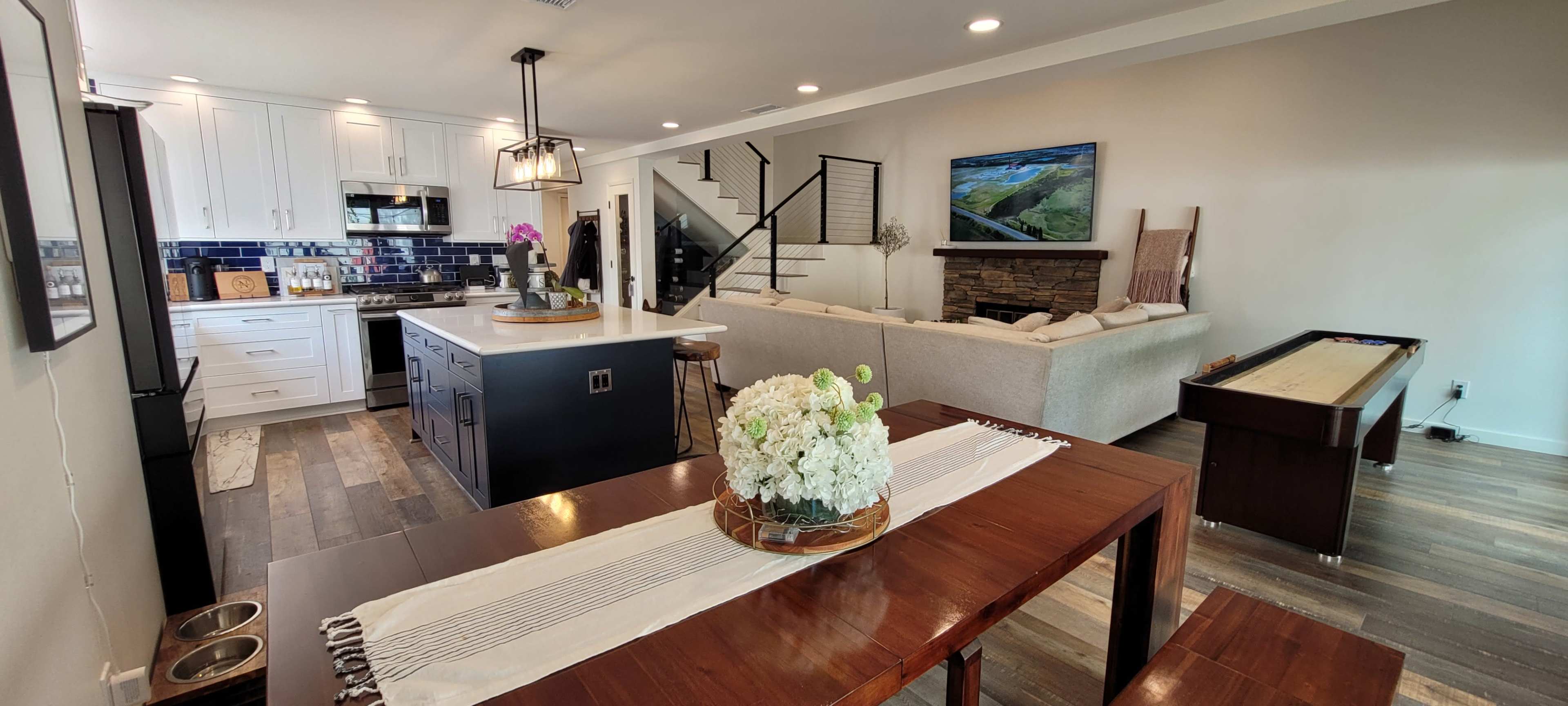 A modern open-concept living and dining area featuring a kitchen with white cabinets, a wooden dining table, and a sectional couch facing a media console.