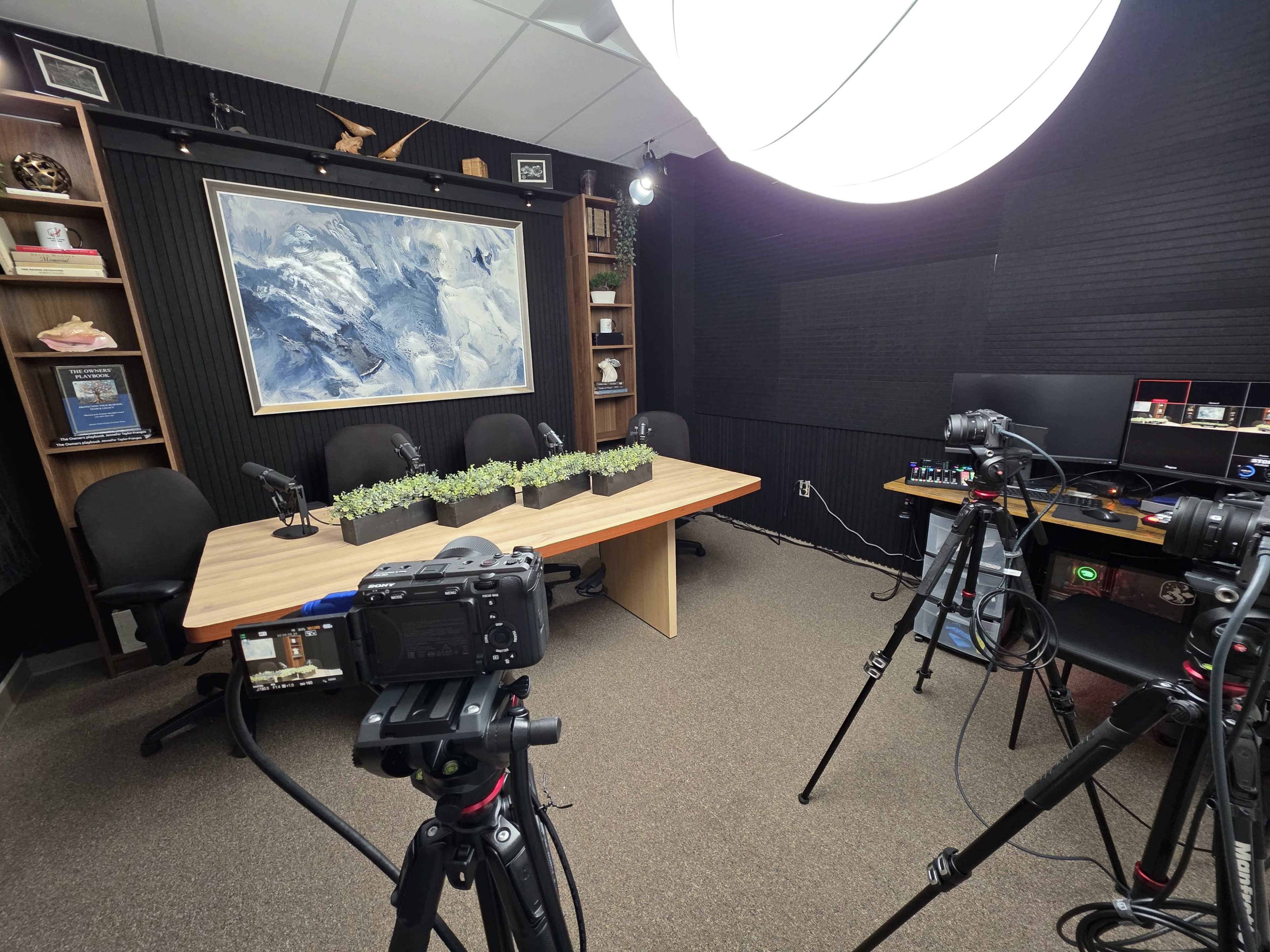 The image shows a media studio with a table set for a discussion, featuring microphones, cameras, and a backdrop of a framed painting.