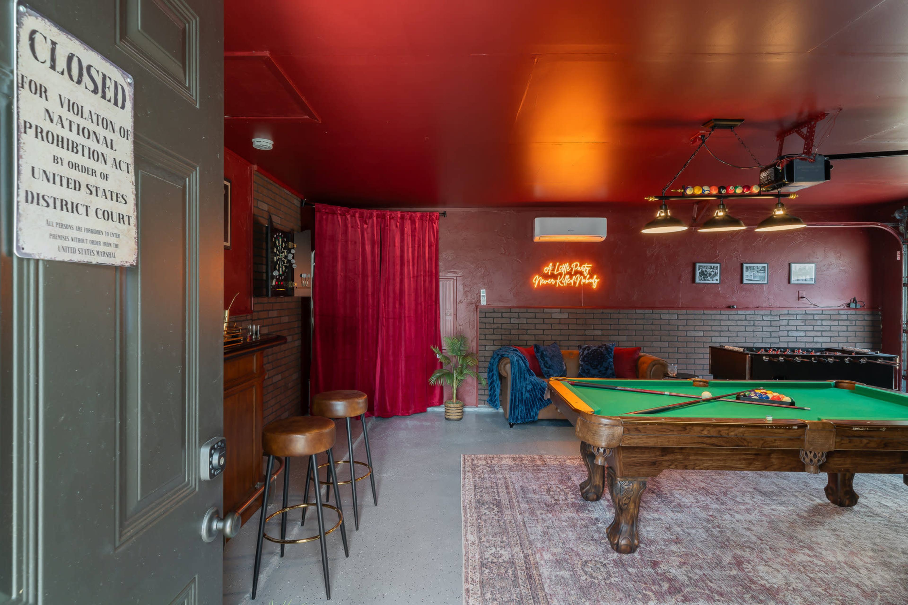 The image shows a recreational room featuring a green pool table, a bar area with stools, and decorative elements including a neon sign and a red curtain.