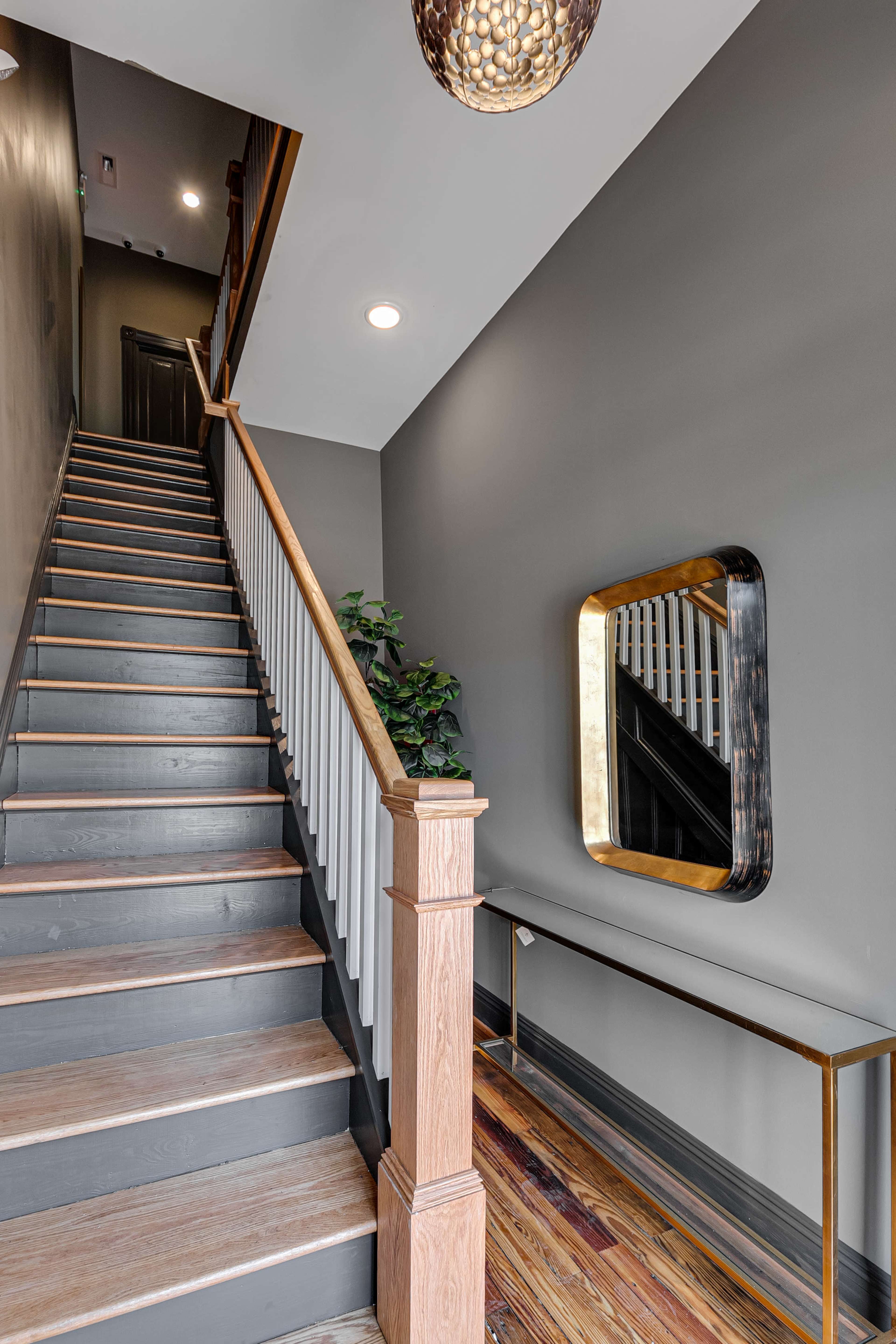 A staircase leads upward in a hallway with gray walls, an indoor plant, and a wall-mounted mirror above a narrow console table.