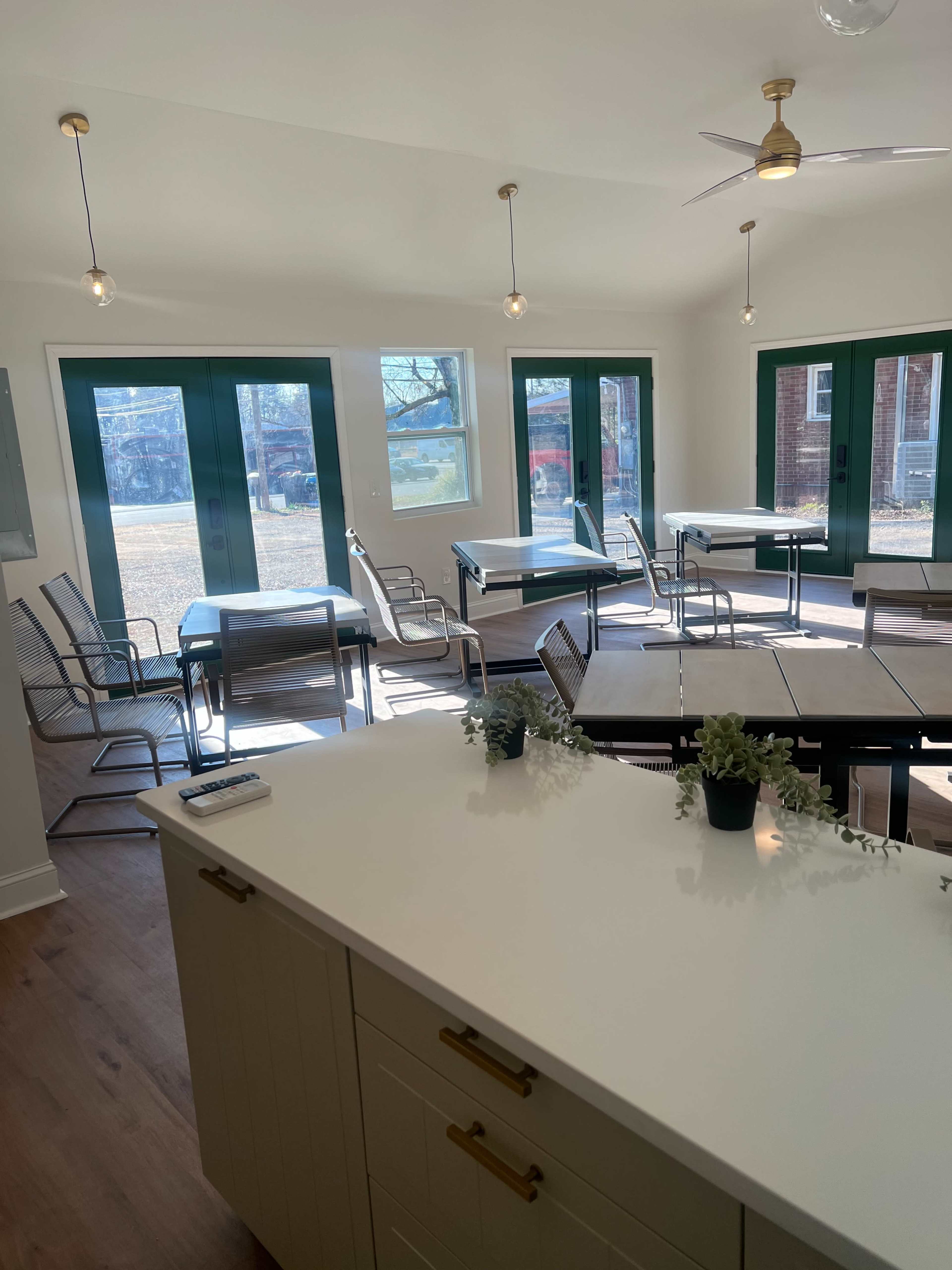 The image shows a bright, well-lit room with several tables and chairs arranged near large green-framed windows.