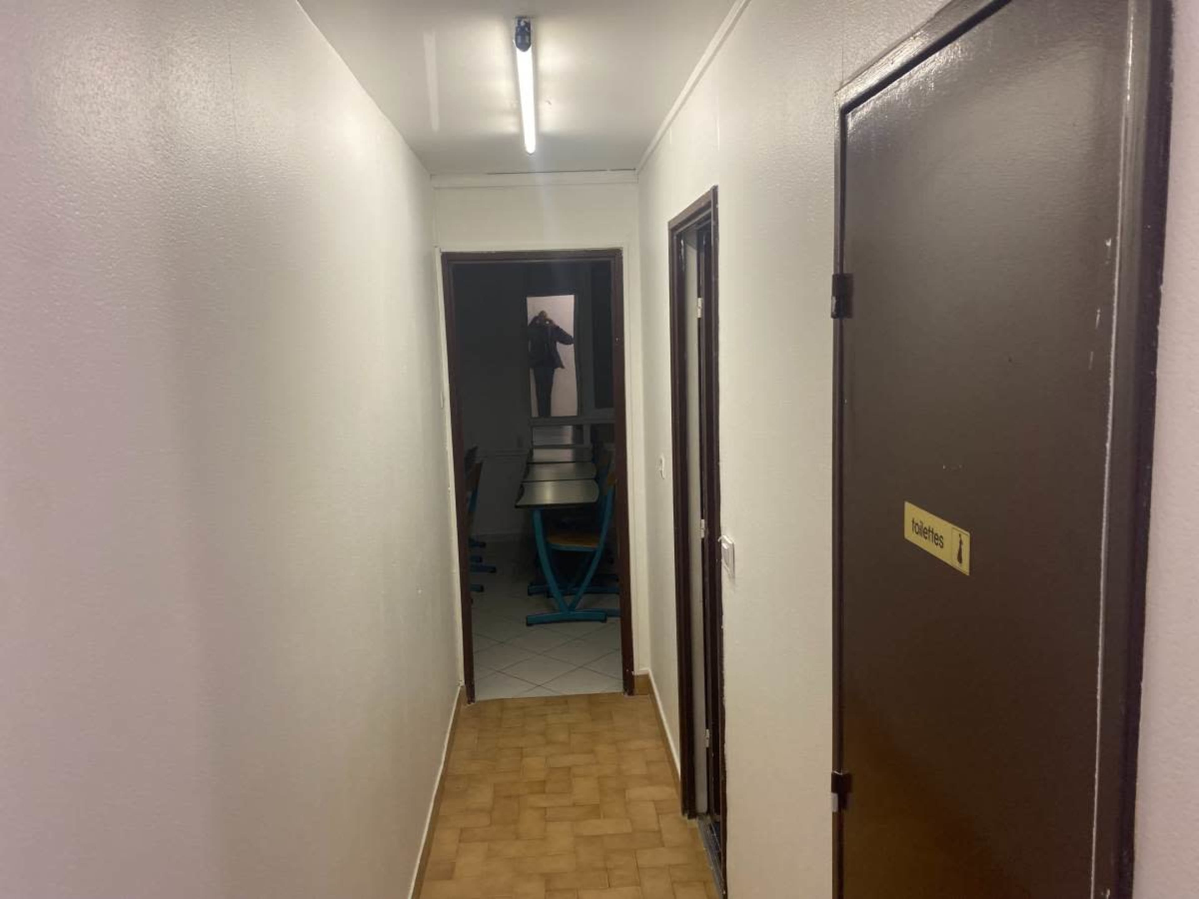 The image shows a narrow, illuminated hallway with doors on either side leading to rooms and a visible table in the distance.