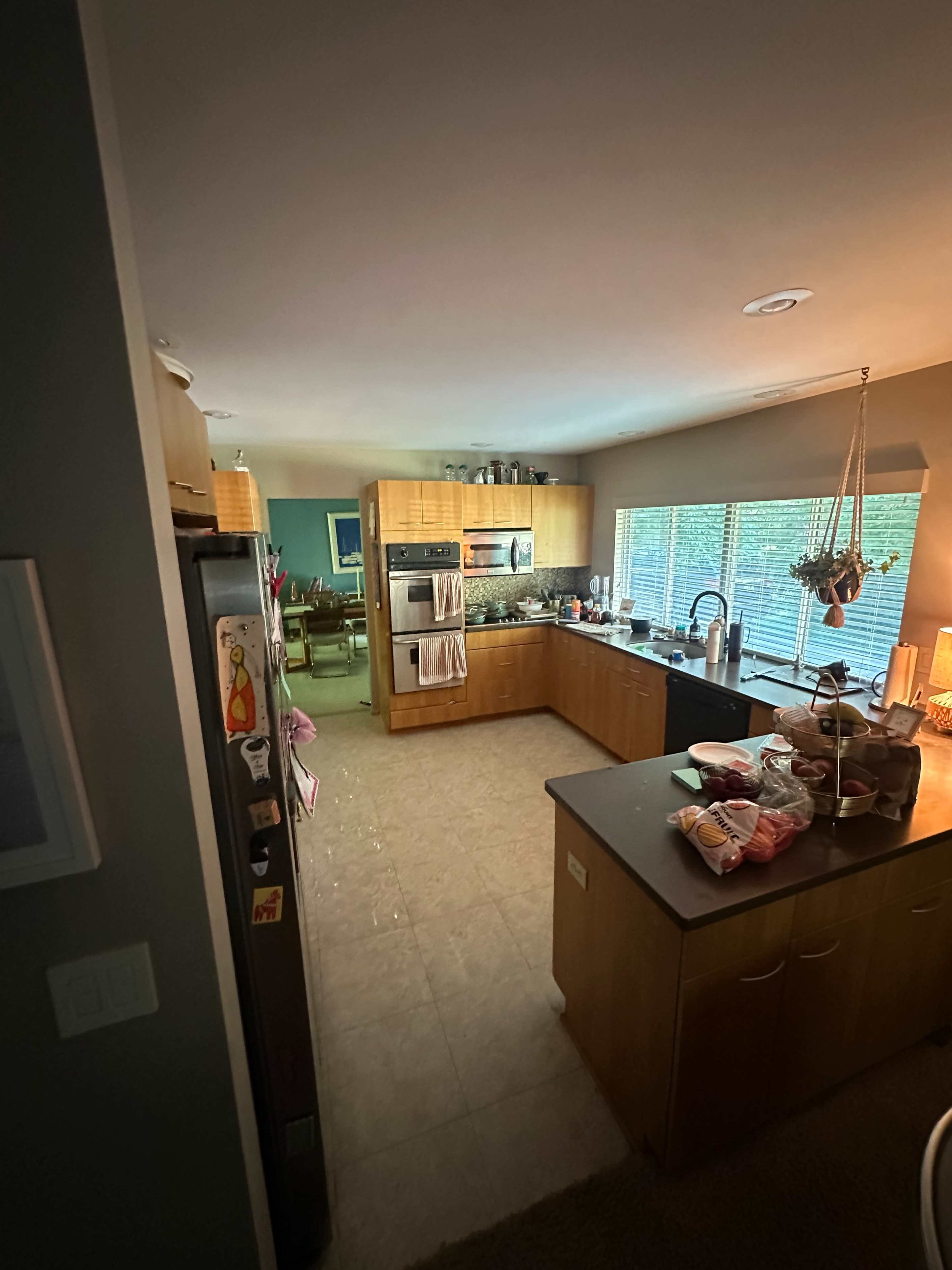 The image shows a modern kitchen with wooden cabinetry, a central island, and large windows allowing natural light to enter the space.