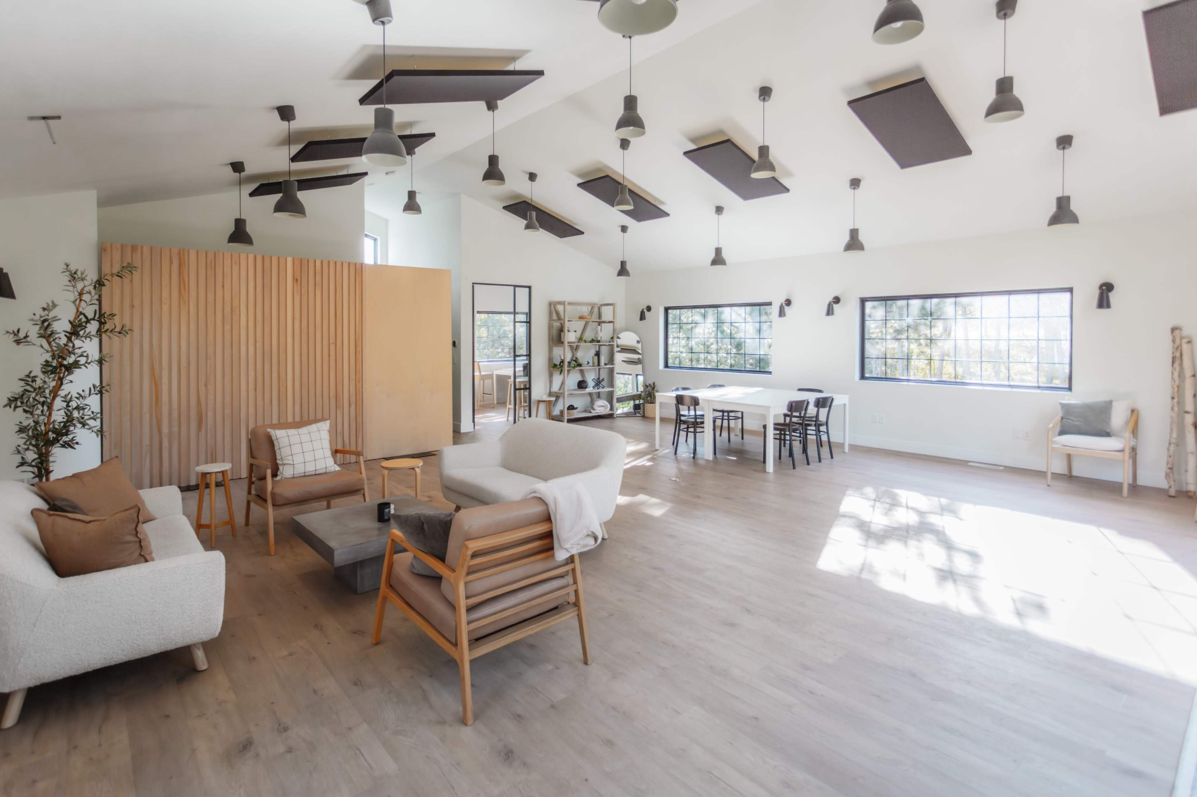 The image shows a spacious, modern interior of a bright room with wooden flooring, featuring a seating area with sofas, a wooden partition, and a dining table.