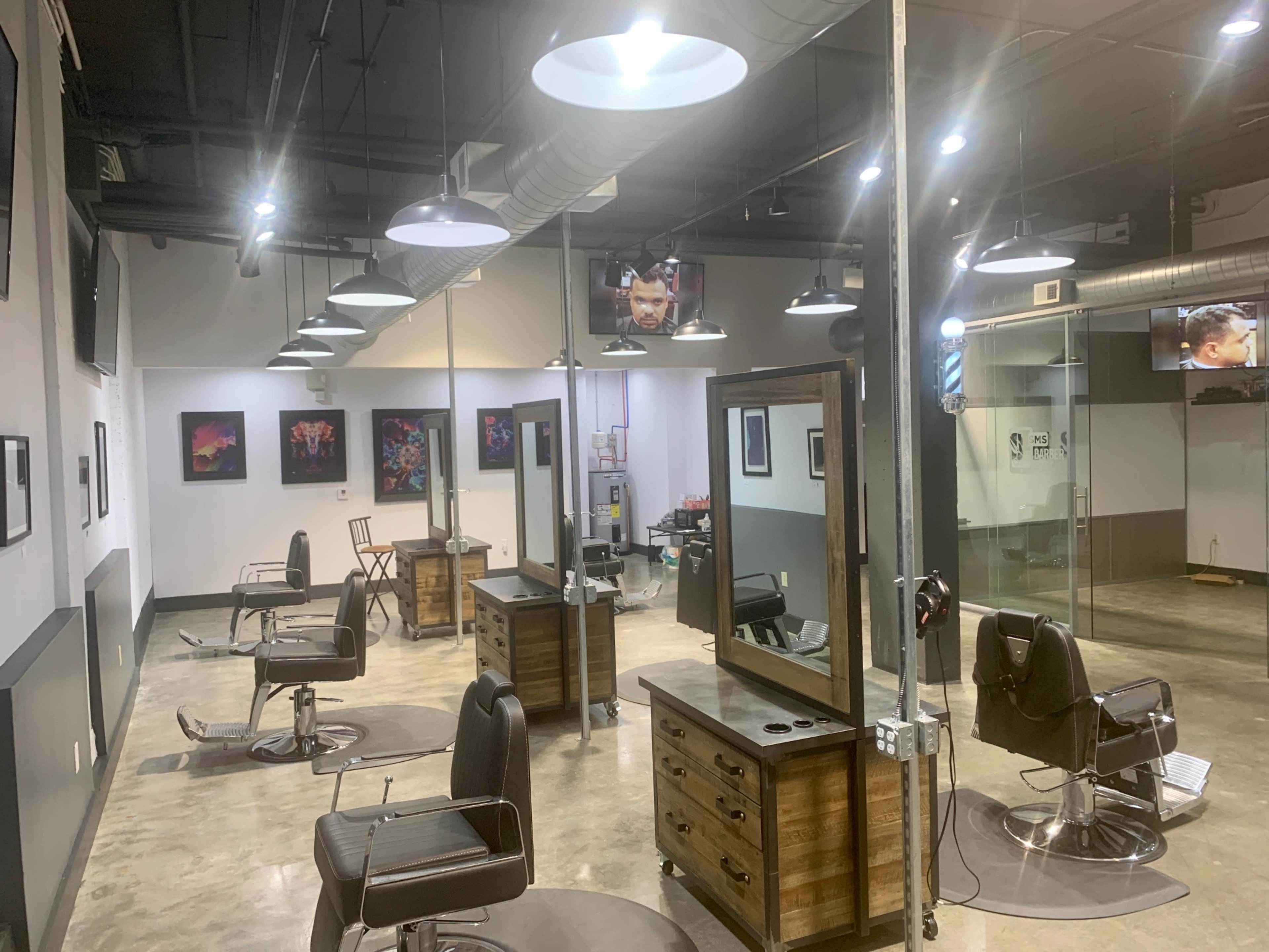 The image shows an empty hair salon featuring several styling stations with mirrors, chairs, and artwork on the walls, illuminated by overhead lights.