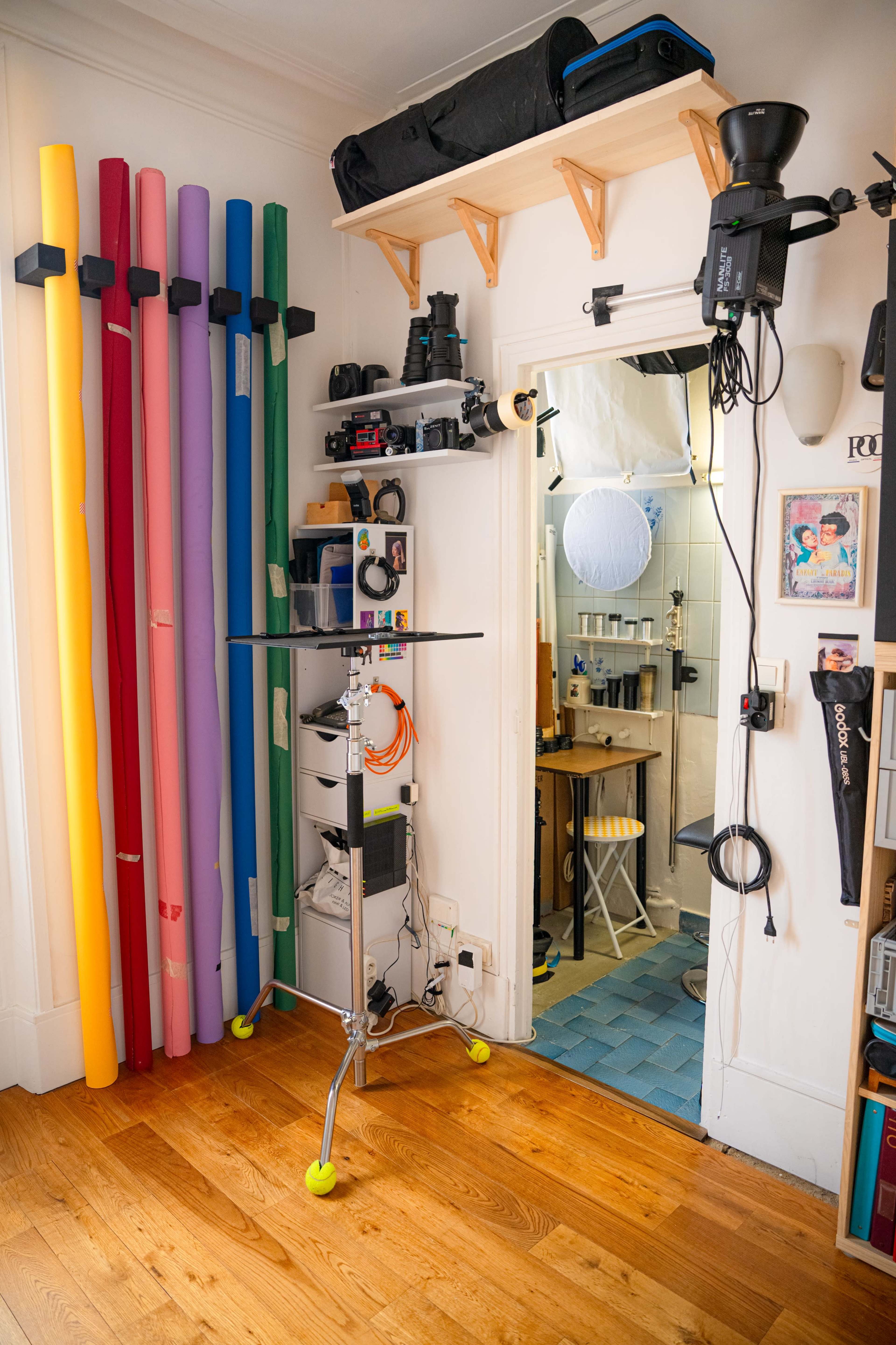 The image shows a photography studio corner featuring various colored backdrop rolls, a camera setup, and a doorway leading to a bathroom.