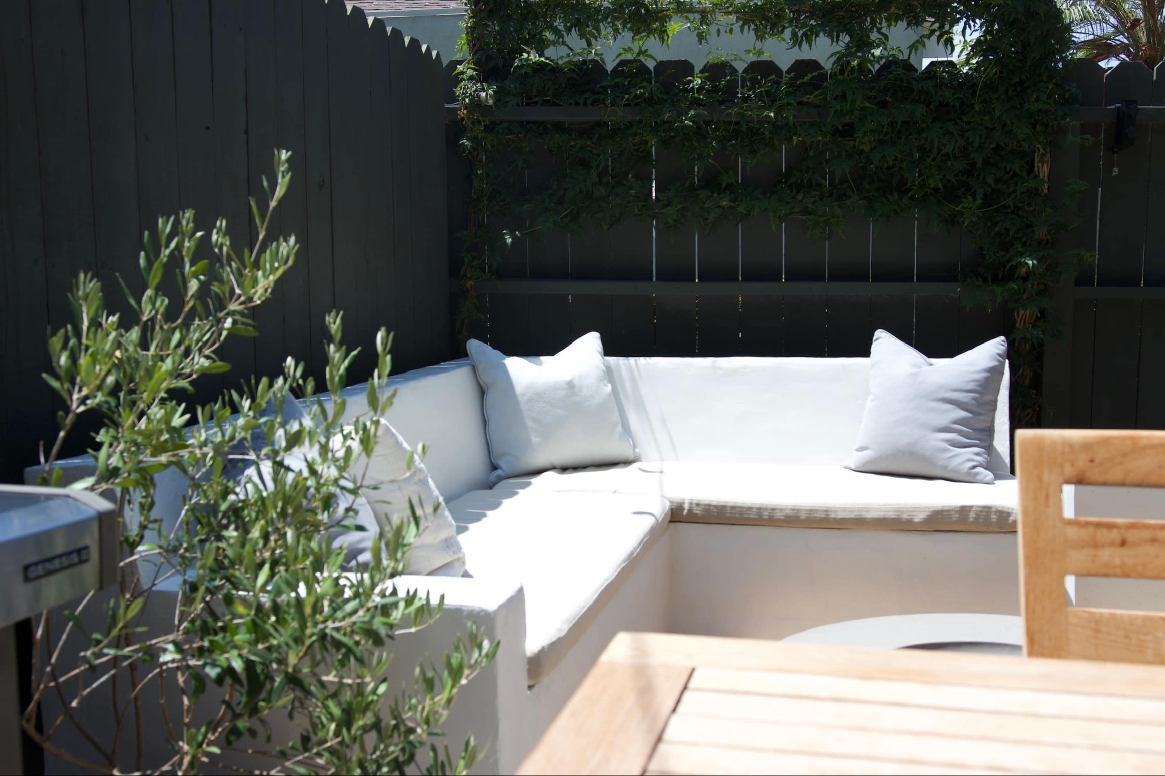 A white outdoor sectional sofa with gray cushions is positioned against a dark wooden fence, surrounded by greenery and a wooden table nearby.