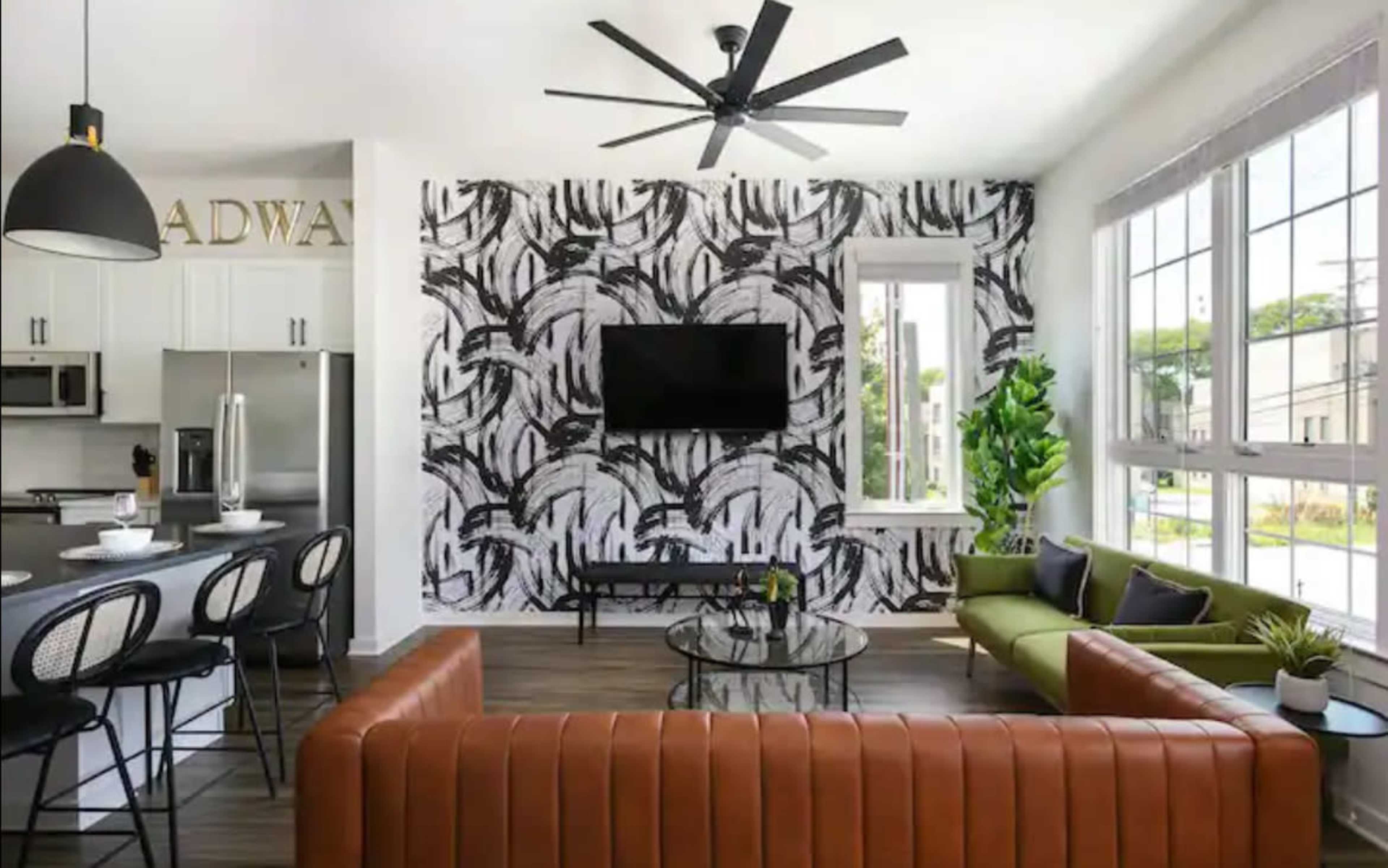 A modern living room featuring a brown leather sofa, a green couch, a black and white patterned accent wall, and a television mounted on the wall, with large windows letting in natural light.