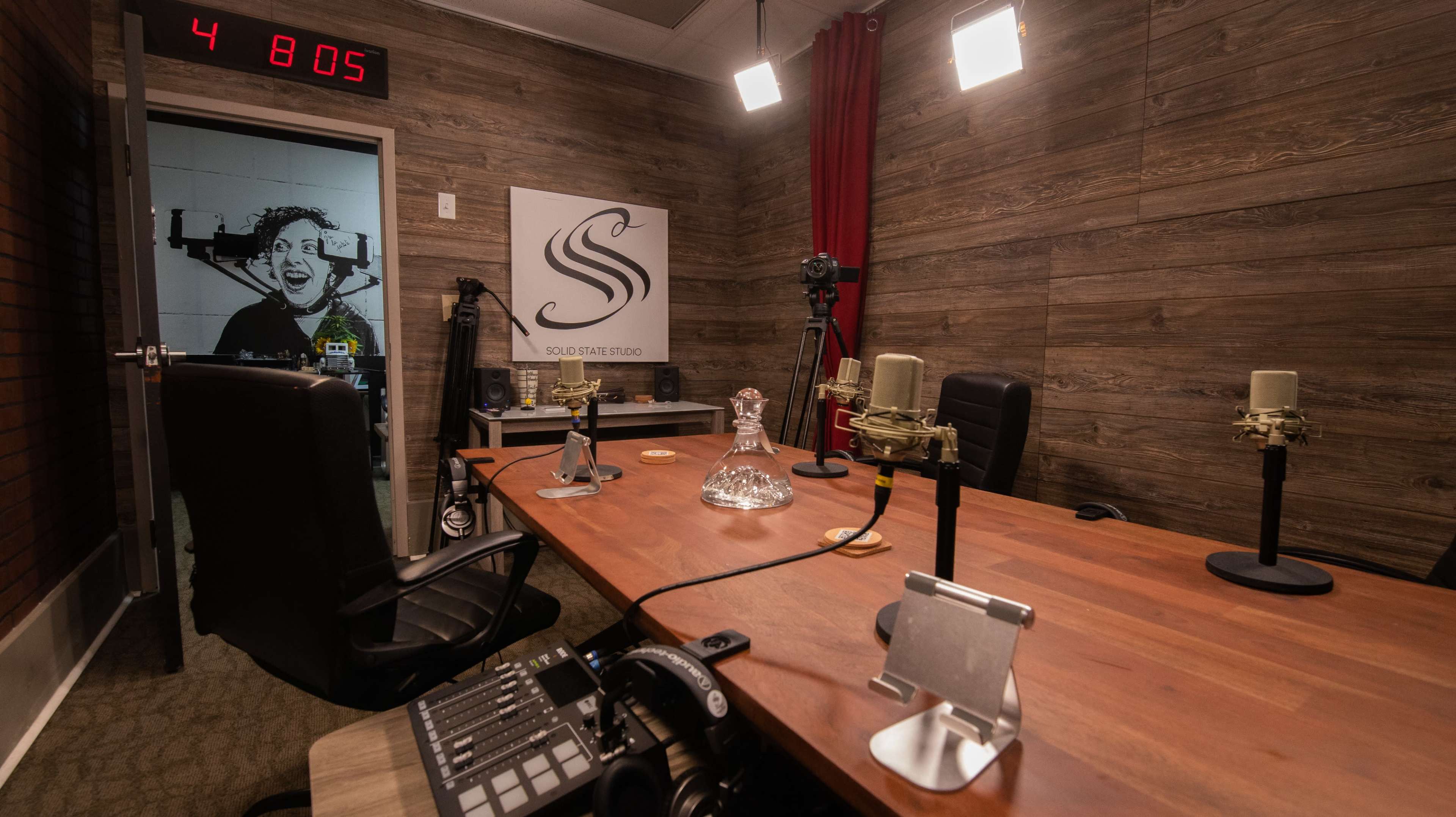 The image shows a recording studio with a wooden table, several microphones, a control panel, and a door featuring a black-and-white photograph.