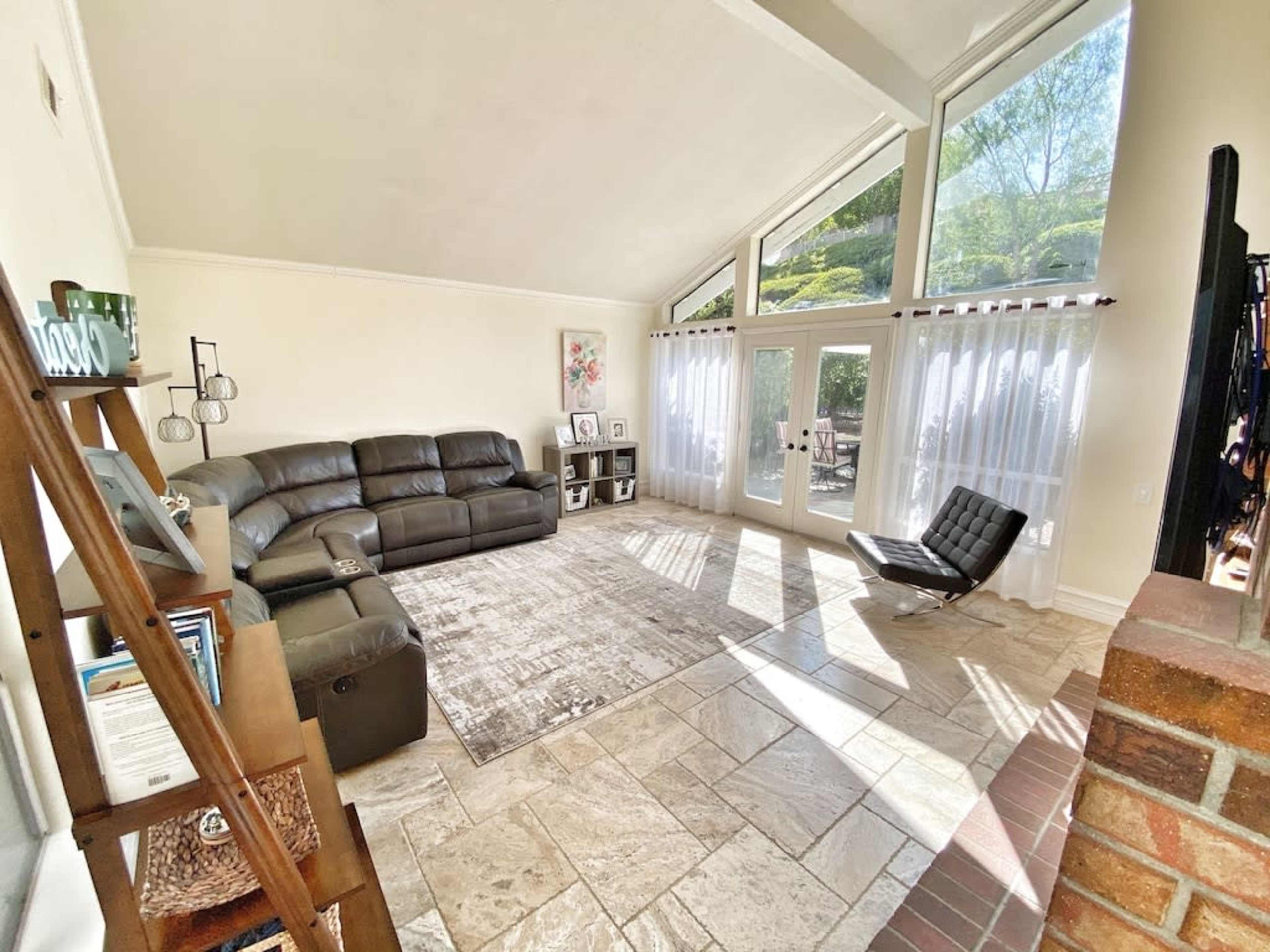 A spacious living room with large windows, a dark leather sectional sofa, and a sleek black lounge chair on a stone-tiled floor.