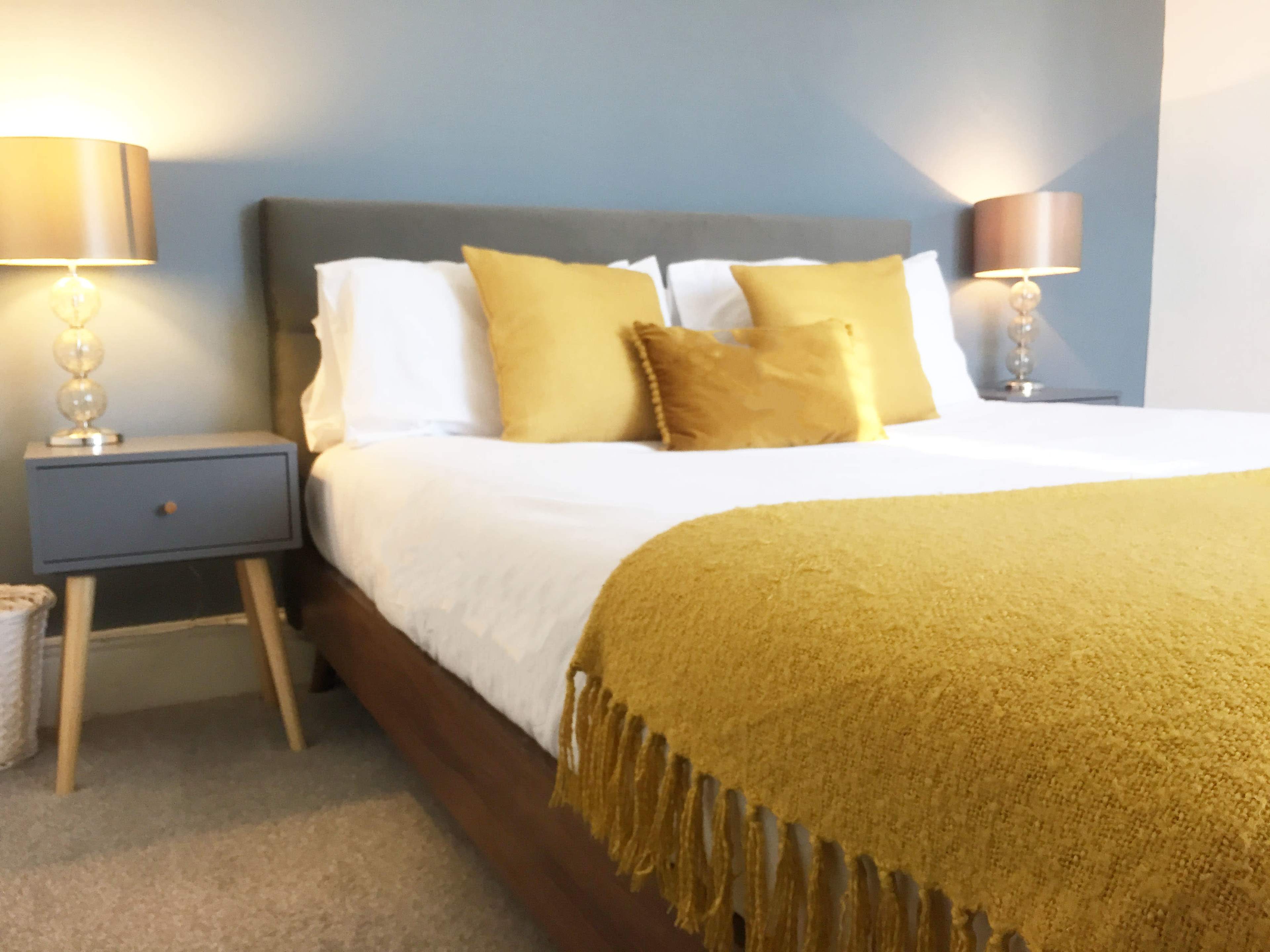 A neatly arranged bedroom featuring a bed with yellow throw pillows and a fringed yellow blanket, flanked by two bedside tables with lamps.