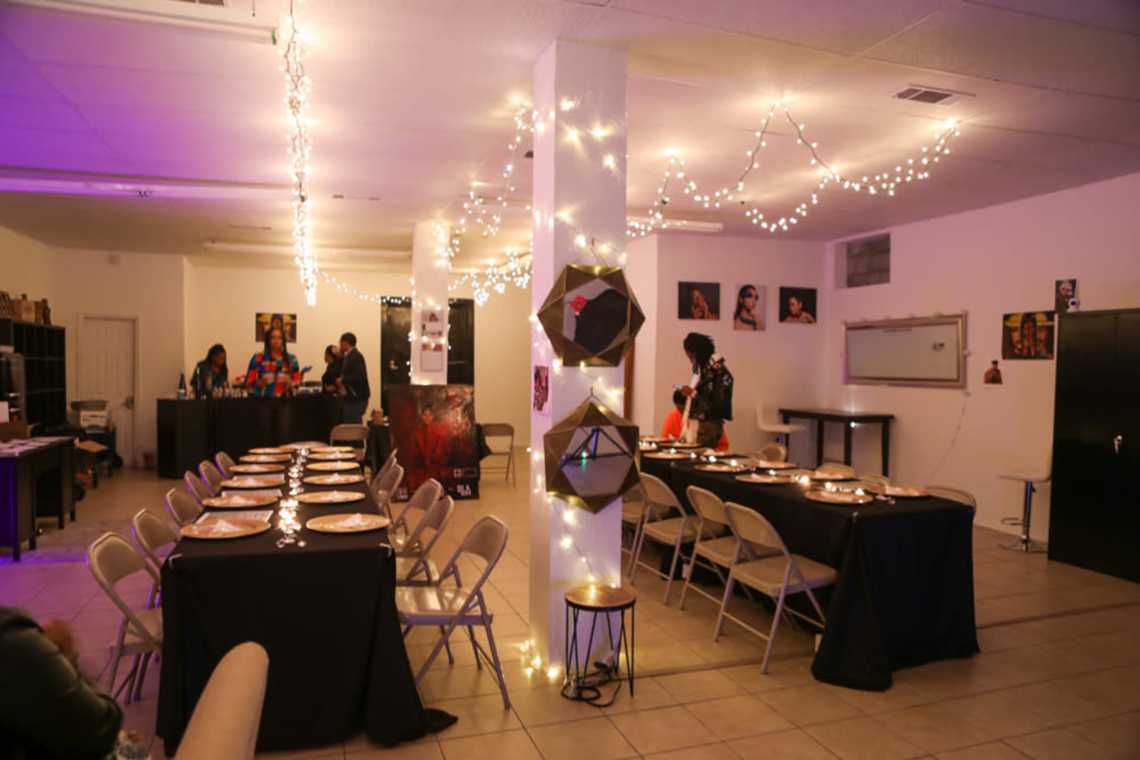 A dimly lit event space with chairs arranged around tables, decorative lights hanging from the ceiling, and a small group of people gathered near a bar area.