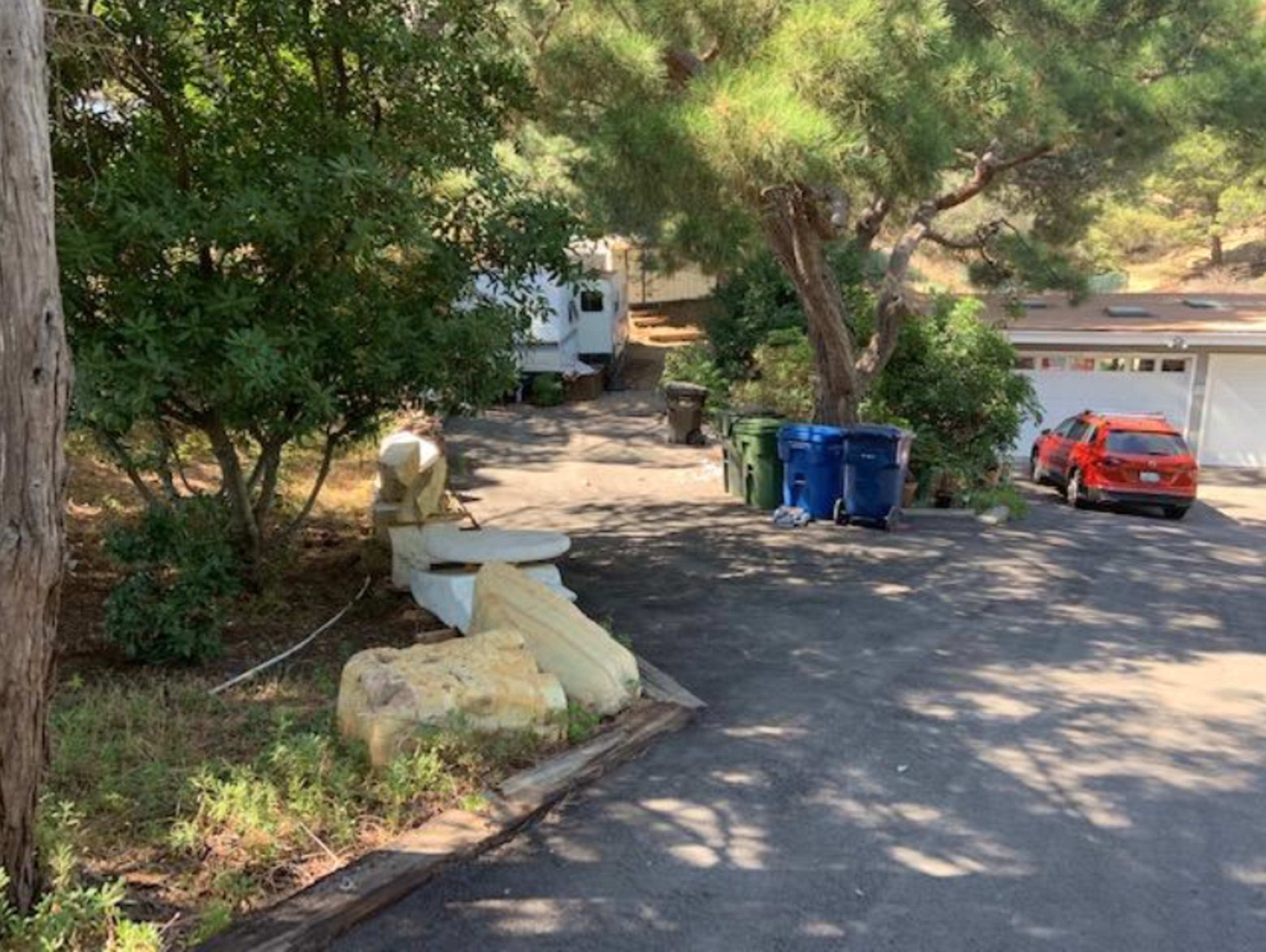 The image shows a sloped driveway leading to a house, with a parked red car near a garage and several trash bins at the side.