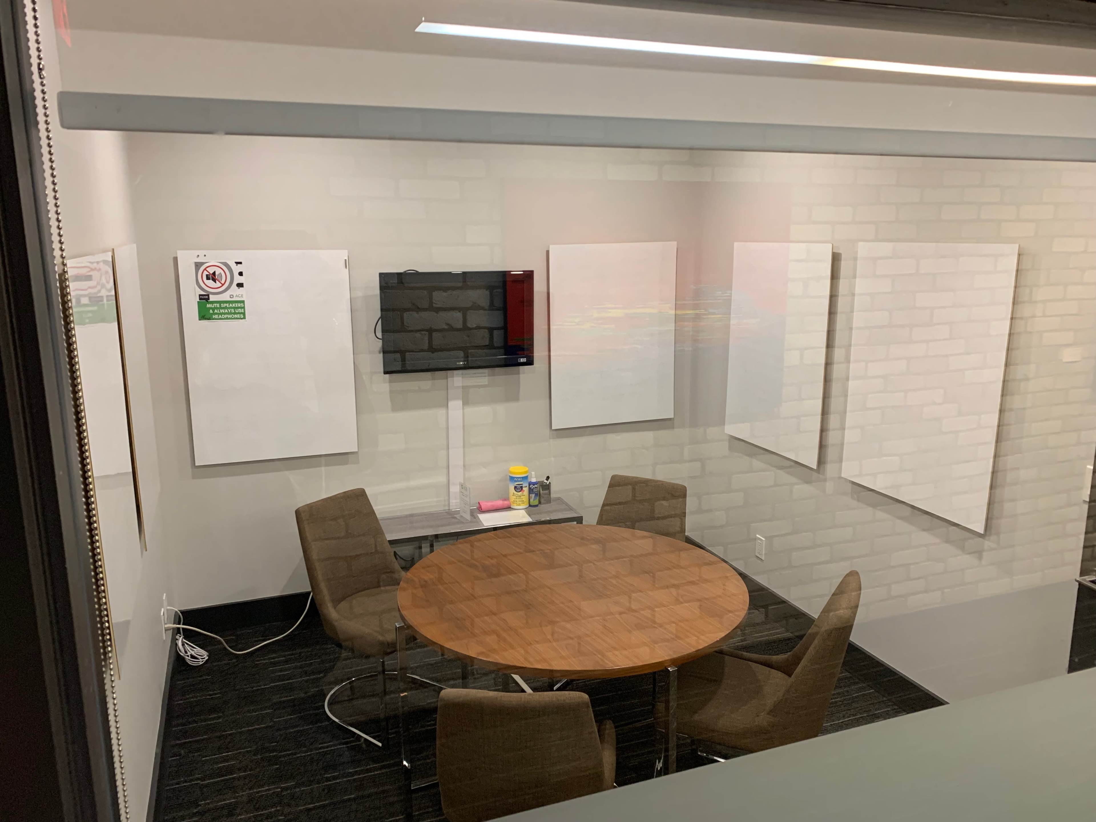 A conference room with a round wooden table surrounded by four chairs, a television mounted on the wall, and several blank whiteboards.