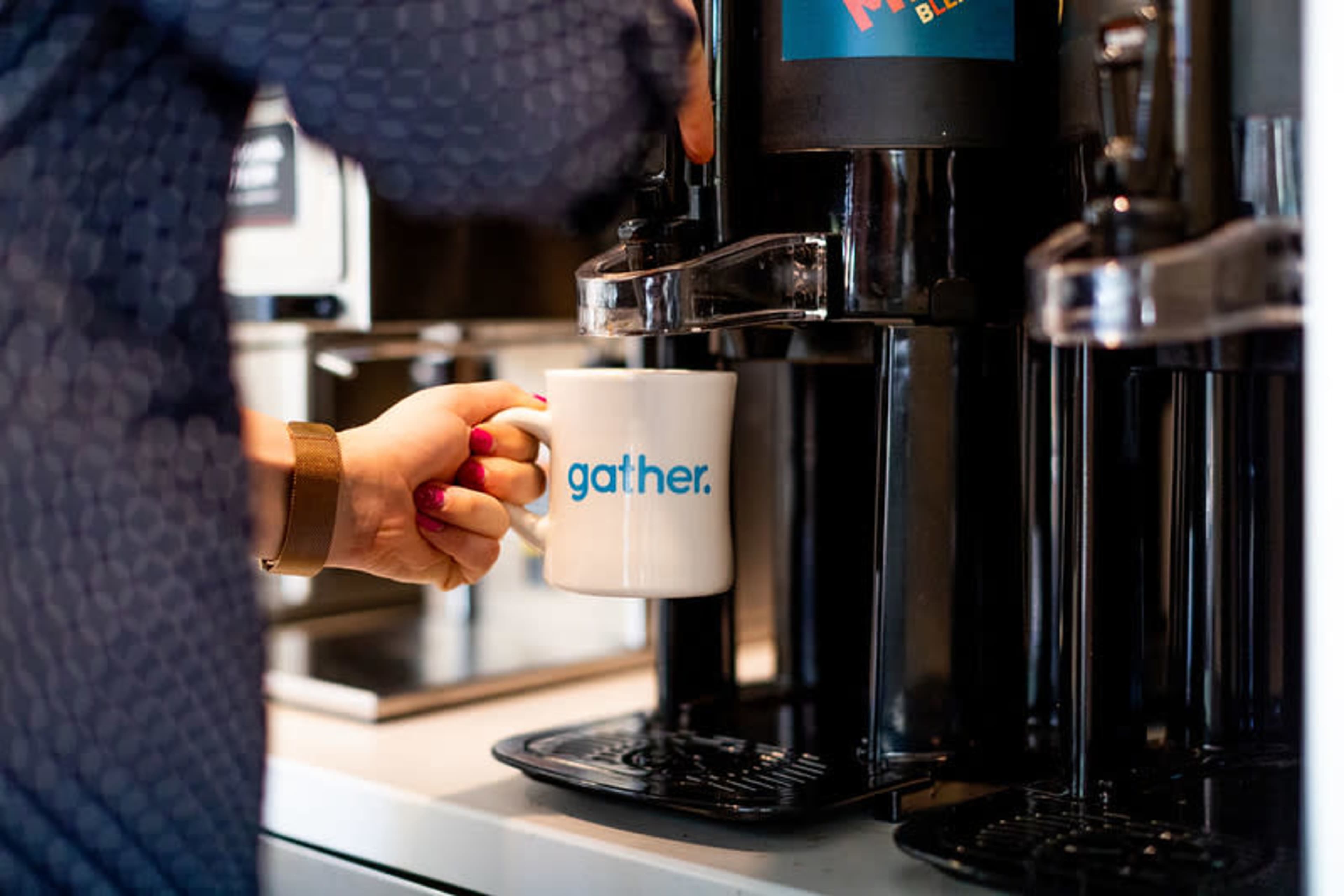A person is pouring coffee into a white mug with the word "gather" printed on it from a coffee machine.