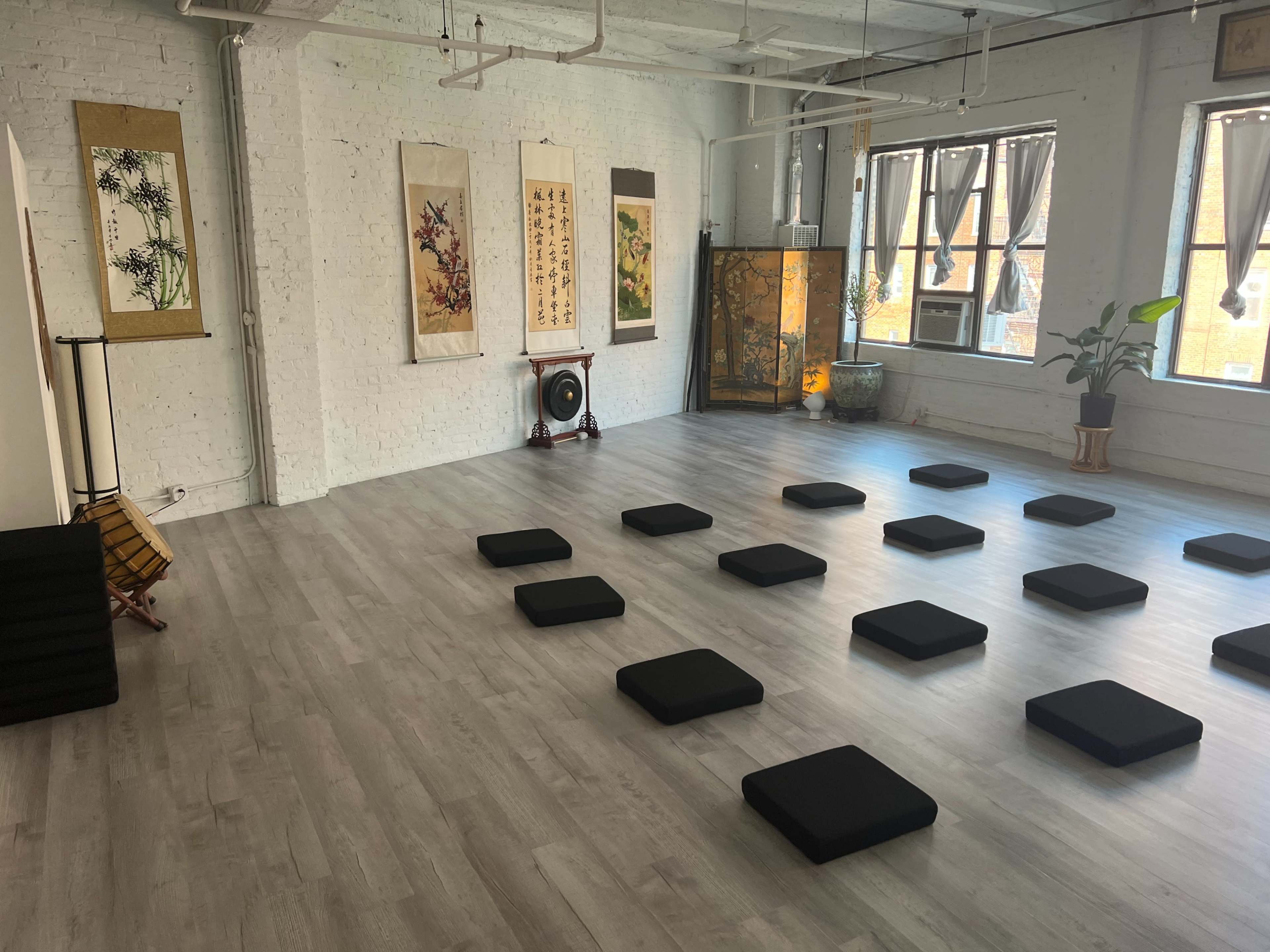 A spacious meditation room features several black cushions arranged on a light wood floor, surrounded by traditional Asian artwork and a plant.