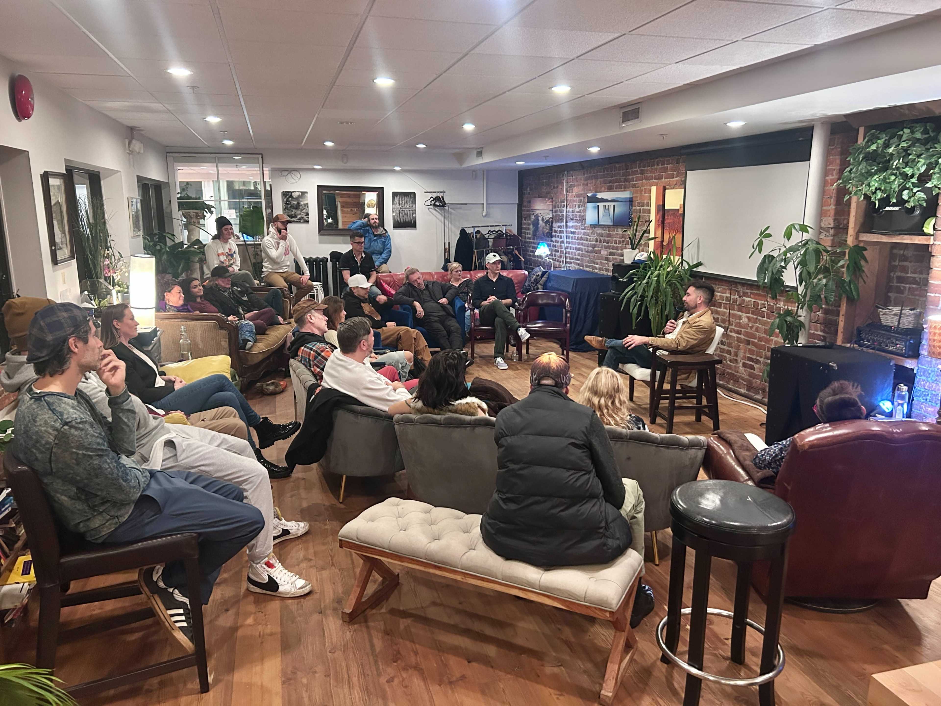 A diverse group of people is seated in a cozy lounge area with exposed brick walls, engaging in a discussion or presentation.