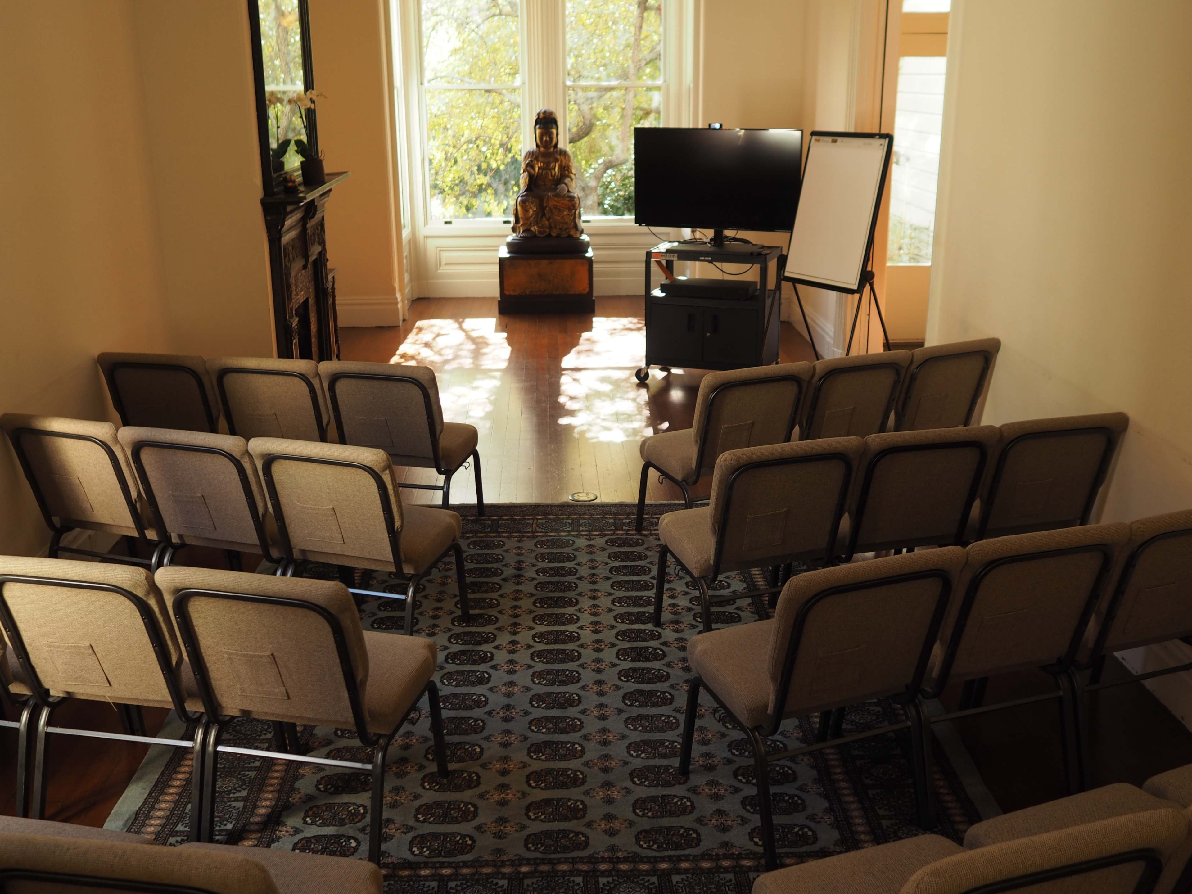 A small seminar room features rows of chairs facing a television and a whiteboard, with a statue of Buddha placed in the corner near a large window.