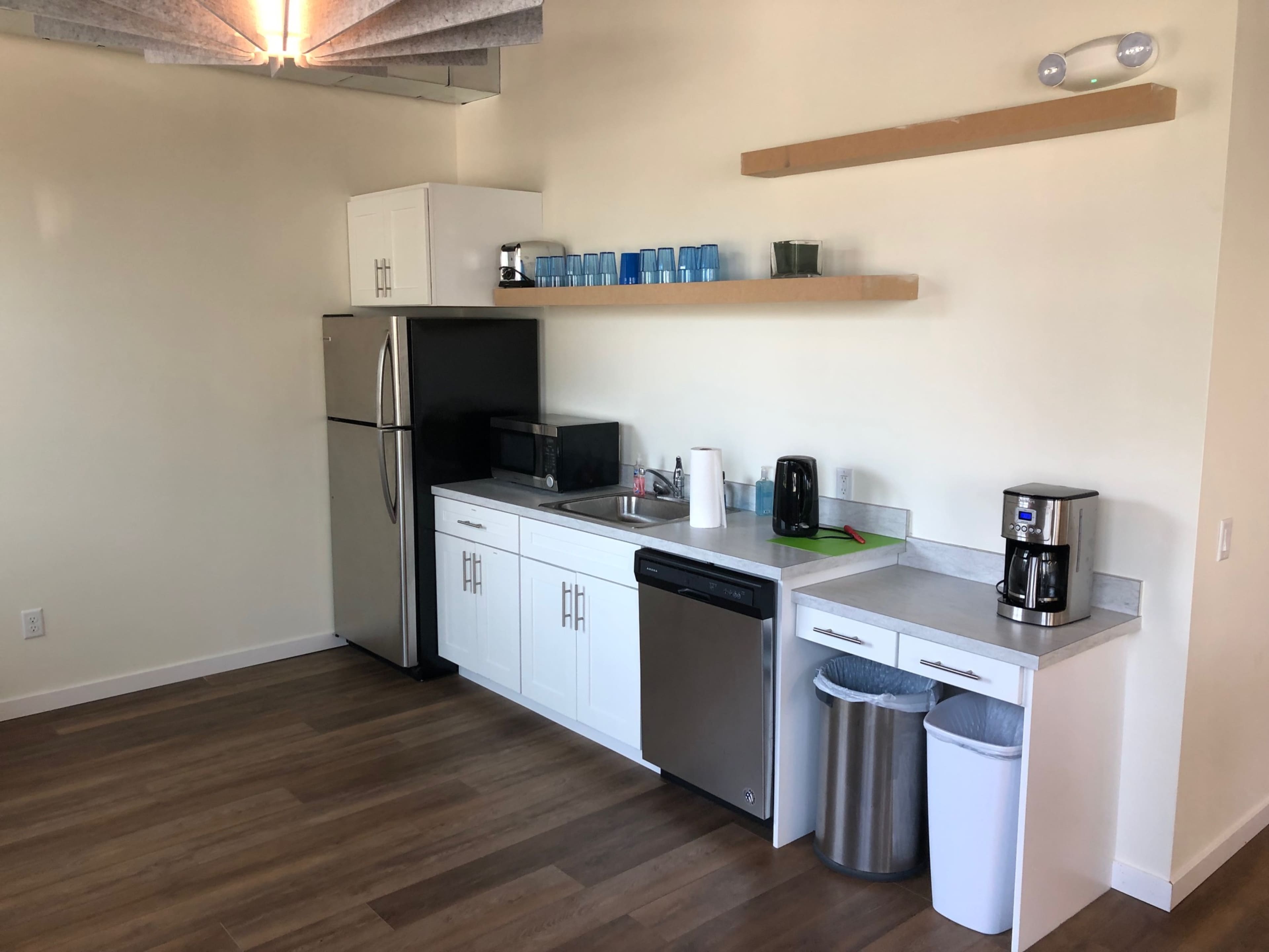 A modern kitchenette featuring a refrigerator, microwave, coffee maker, and a sink with white cabinetry and a wooden shelf above stocked with blue cups.