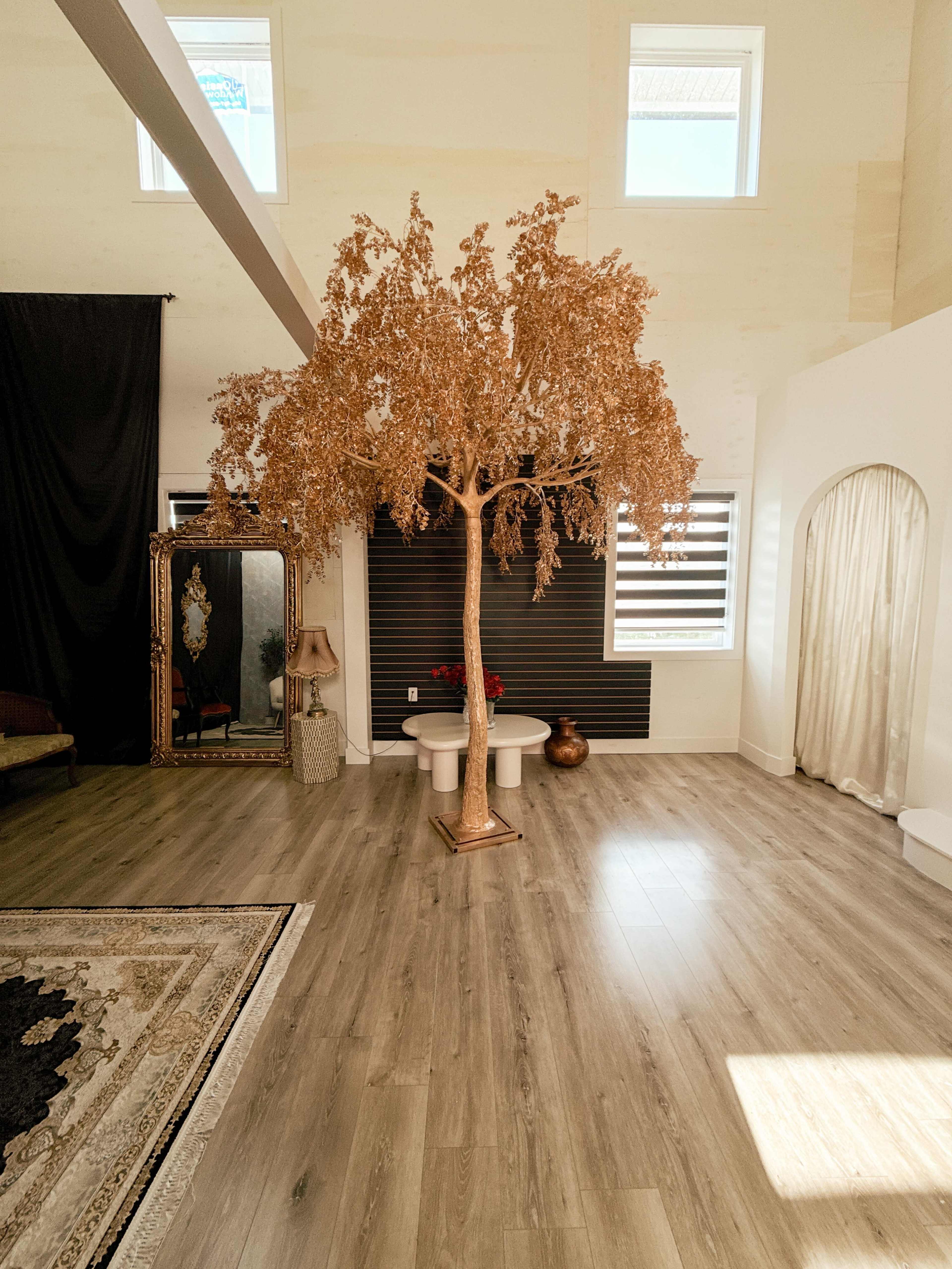 The image shows a spacious room featuring a large artificial tree, a vintage mirror, and a pair of white stools on a wooden floor.