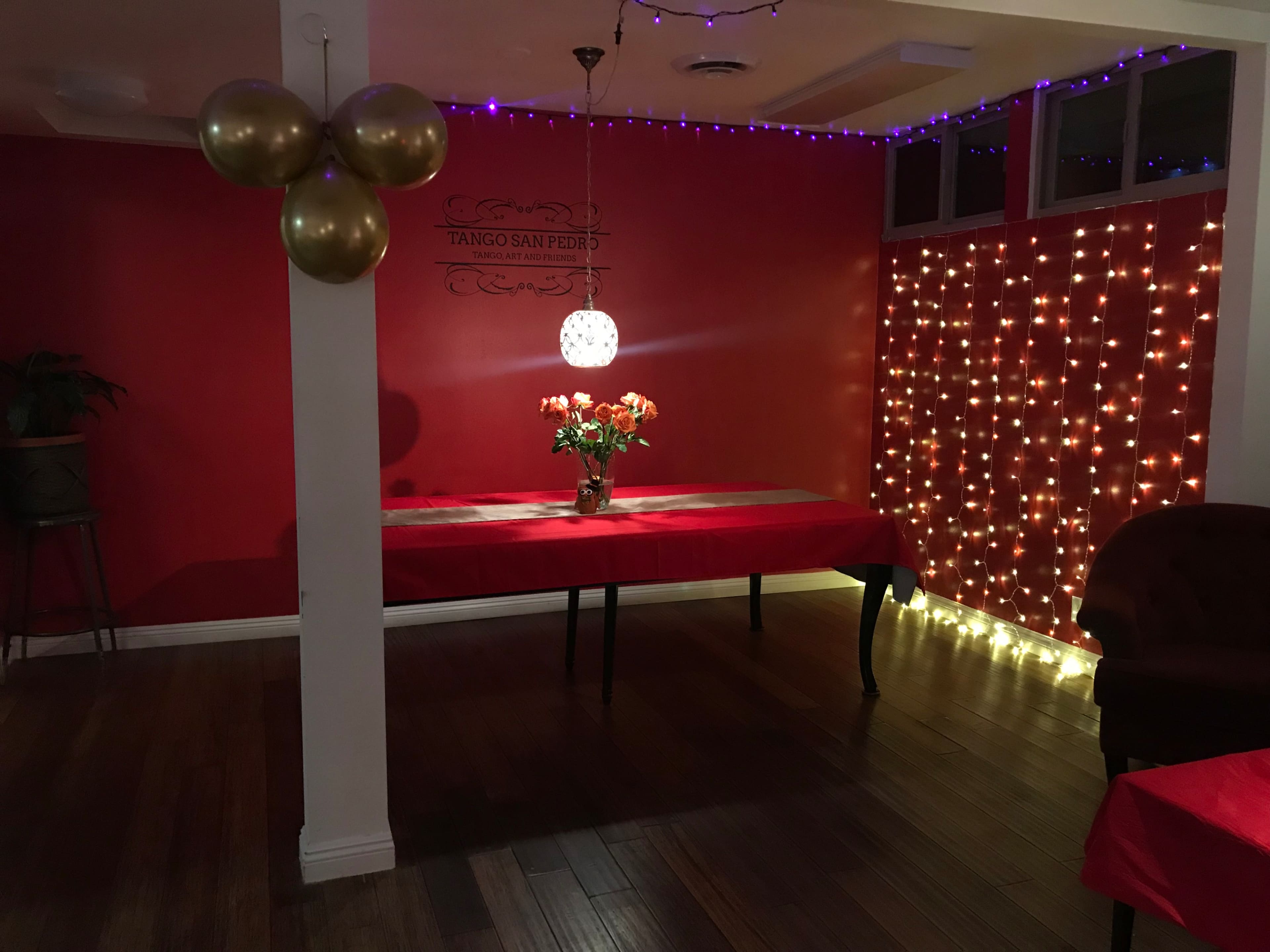 A room with red walls features a table covered in a red cloth, a vase of roses on it, and an illuminated backdrop of string lights.
