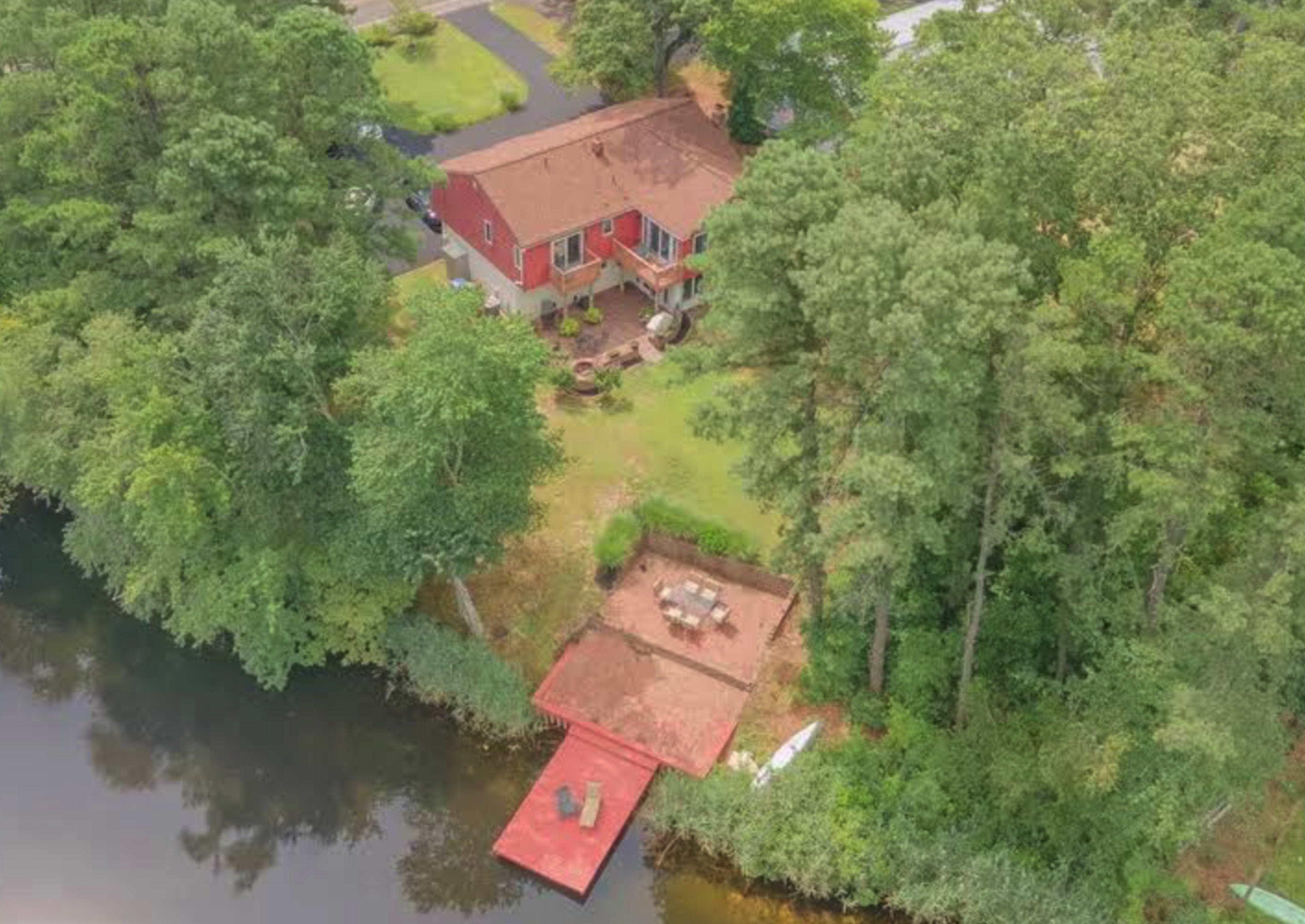 An aerial view shows a red house beside a calm body of water, with a wooden deck and outdoor seating area surrounded by trees.