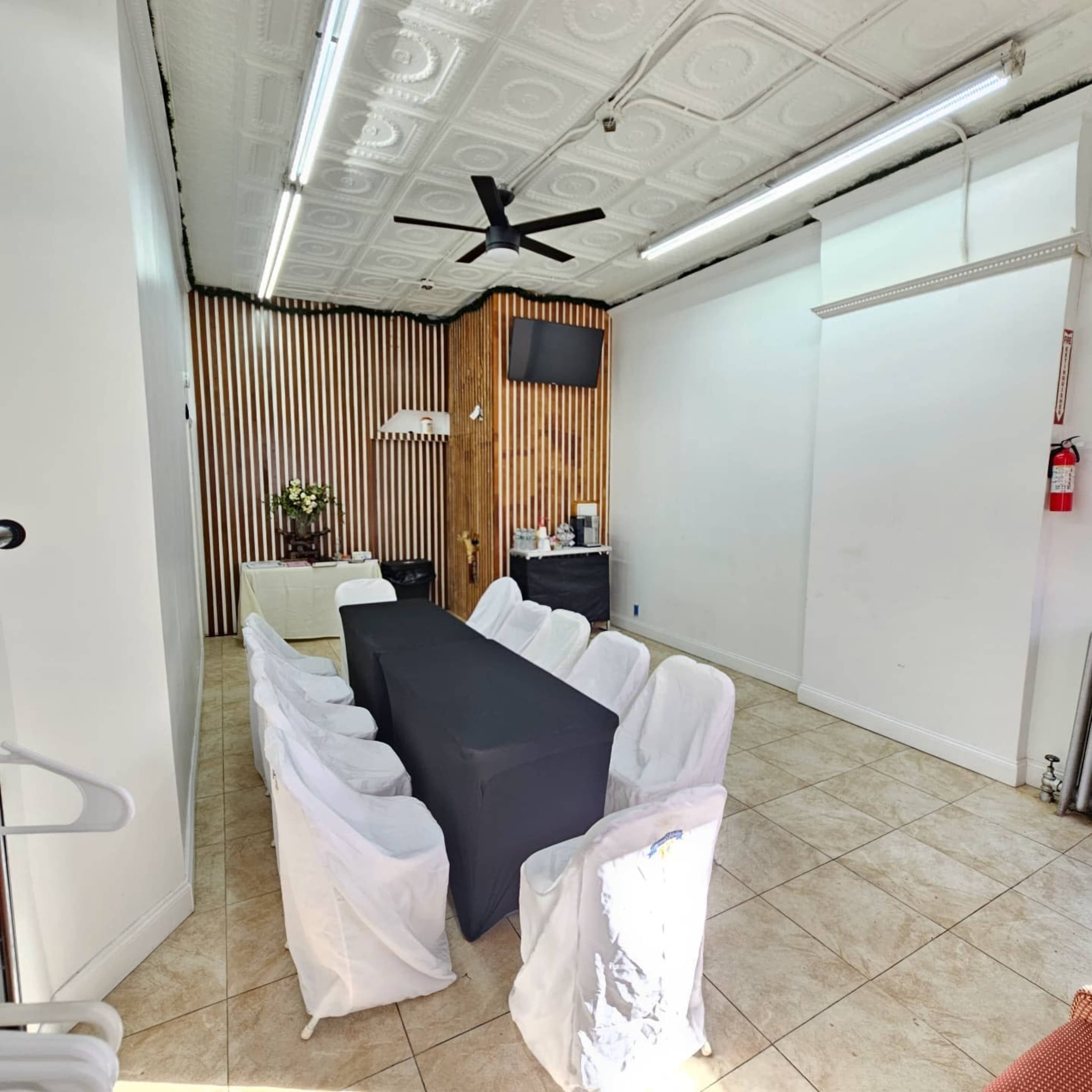 The image shows a tidy meeting room with a long black table surrounded by white chairs, set against a backdrop of wooden paneling and a wall-mounted TV.