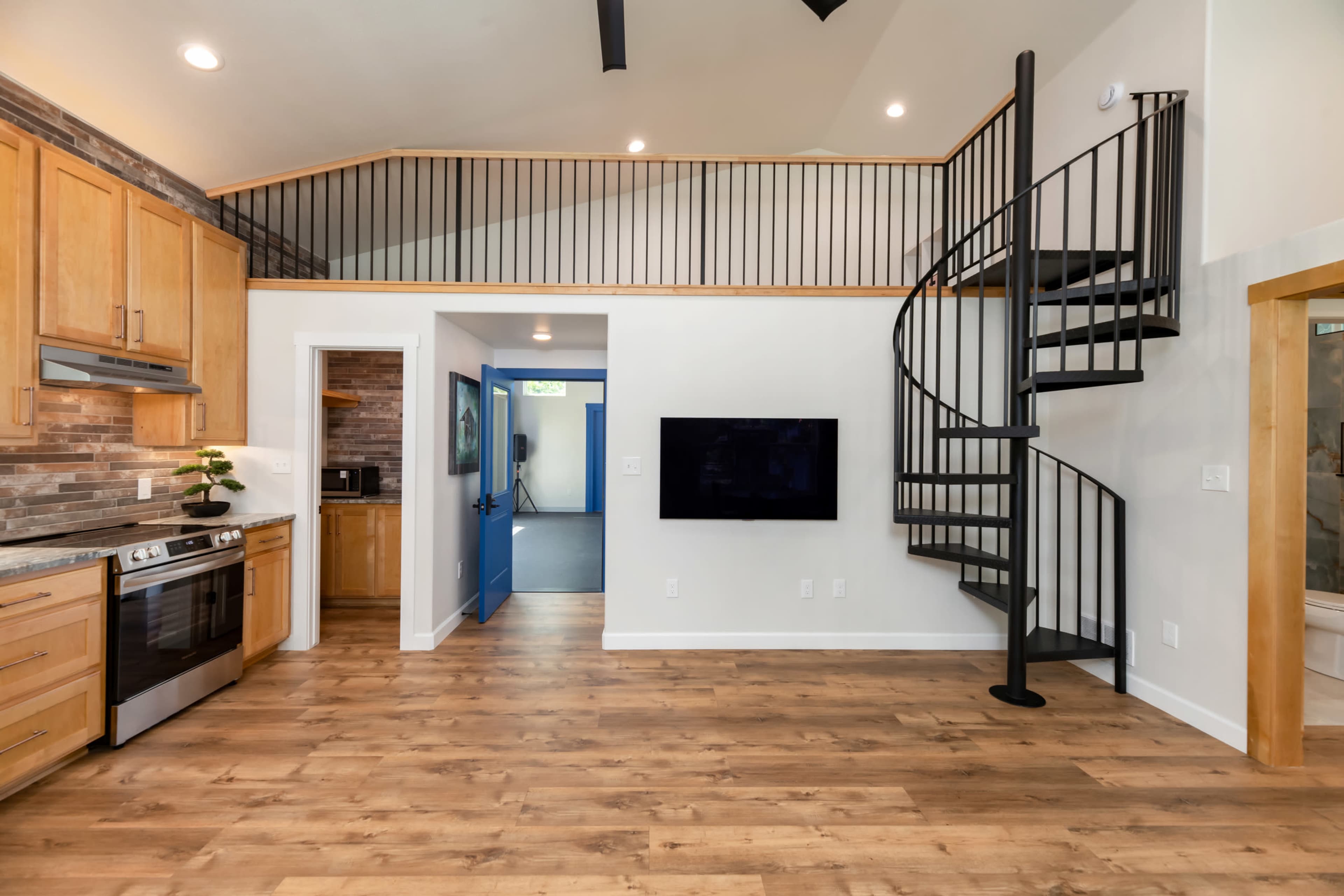 The image shows a modern kitchen and living area featuring wooden cabinets, a spiral staircase, and a large wall-mounted TV.
