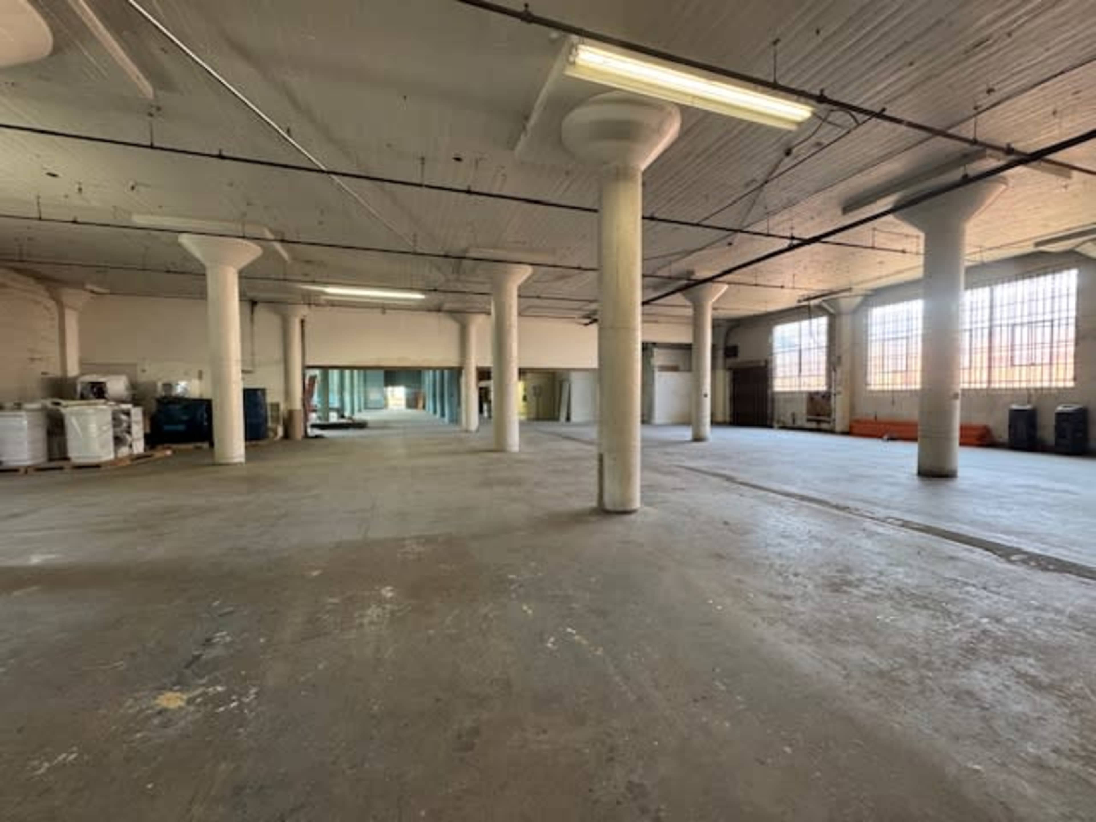The image shows a large, empty industrial space with concrete floors and several columns, illuminated by natural light through tall windows.