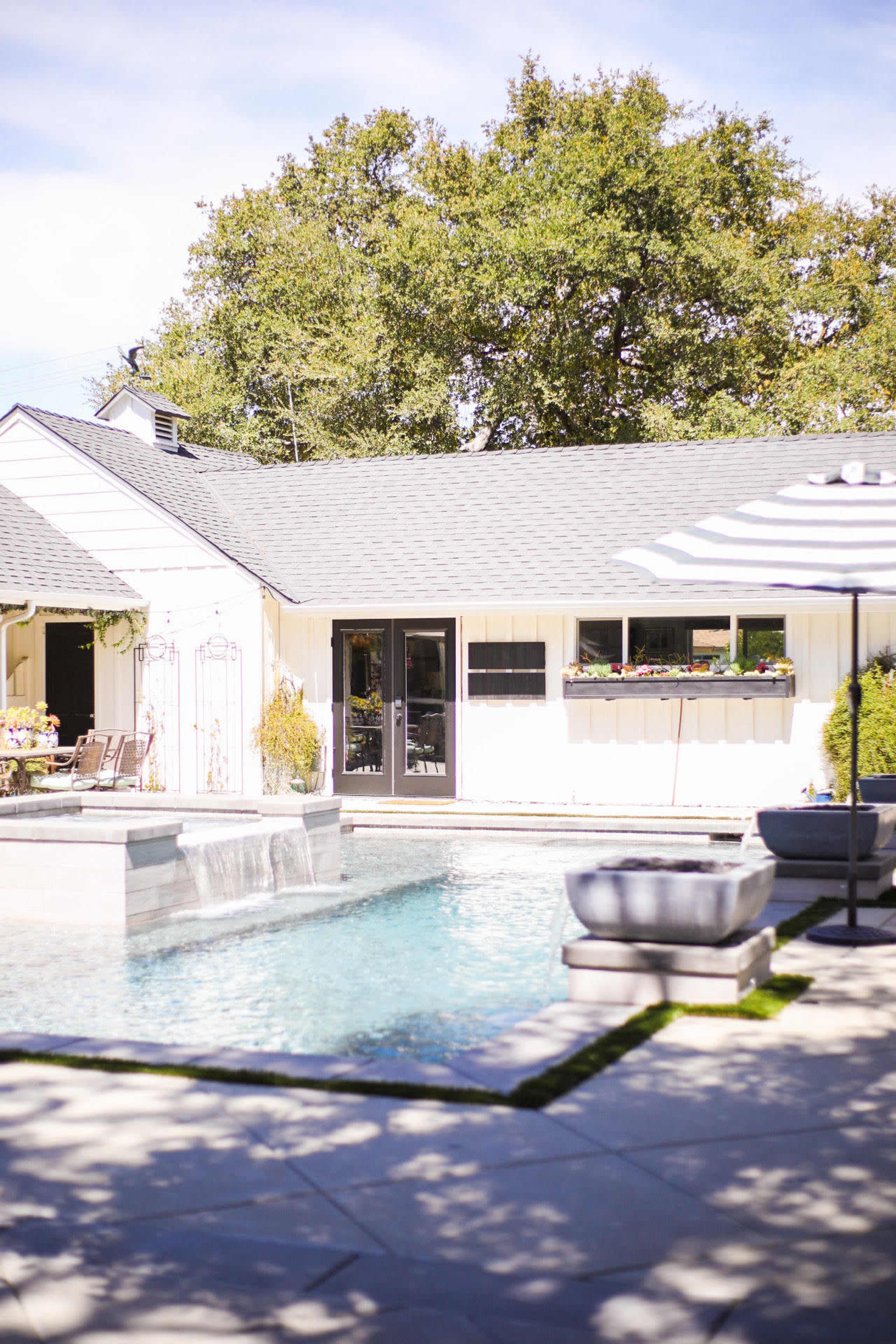 A modern backyard features a swimming pool with a waterfall, surrounded by a house, patio furniture, and lush greenery.