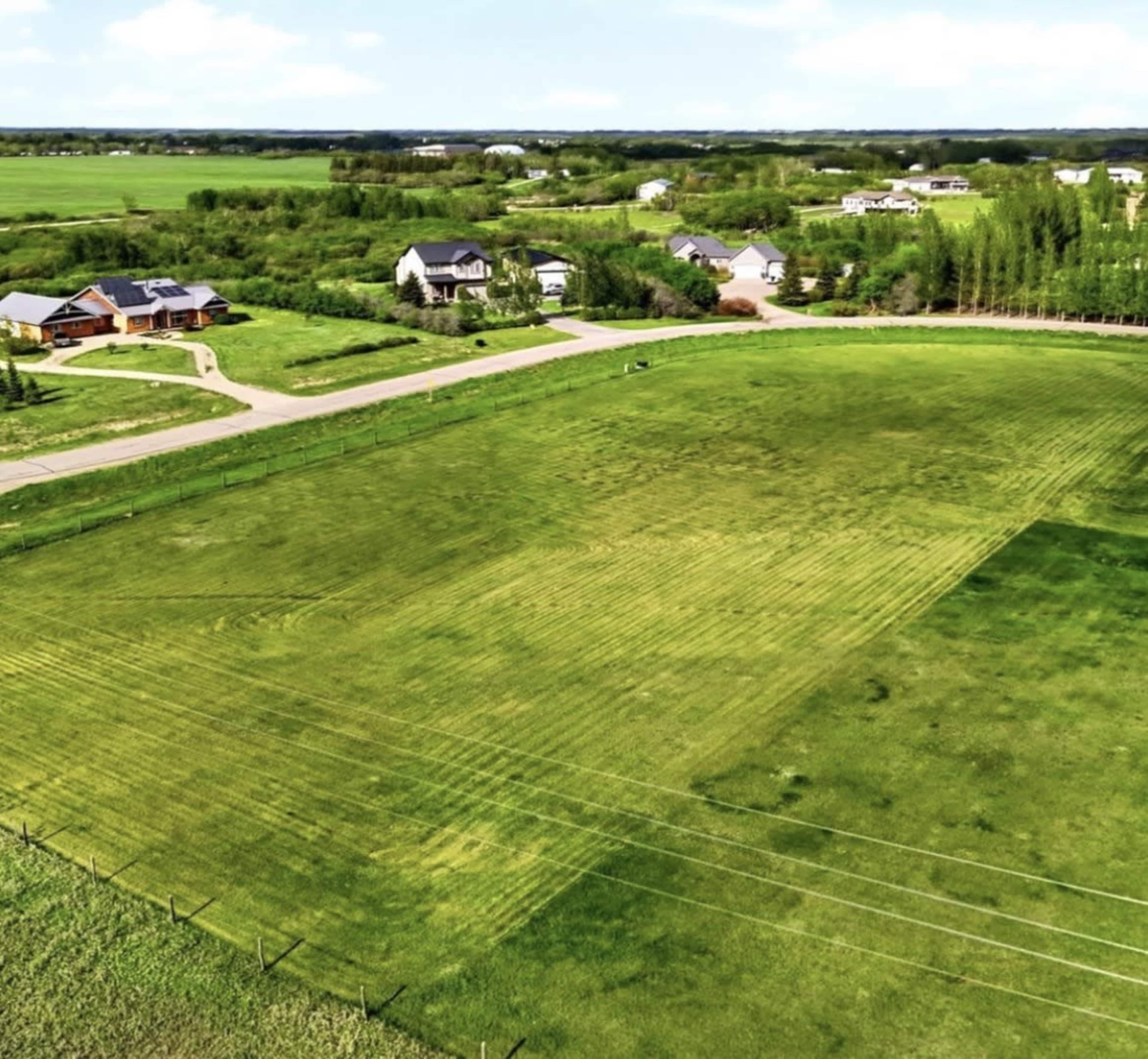An aerial view shows a green field bordered by a winding road and several homes in a rural landscape.