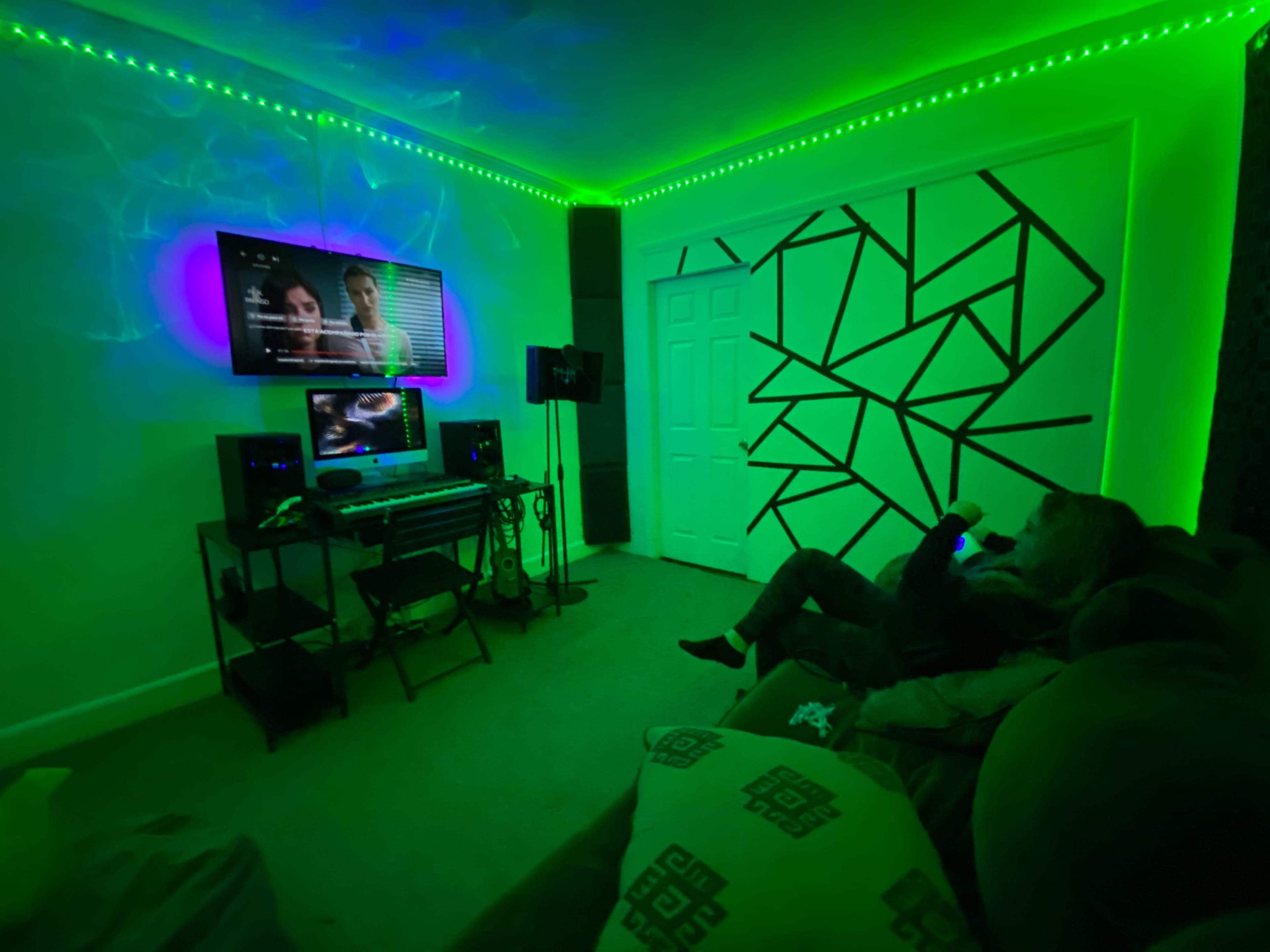 A person sits on a couch in a room illuminated by green LED lights, watching a screen mounted on the wall while various audio equipment is visible nearby.