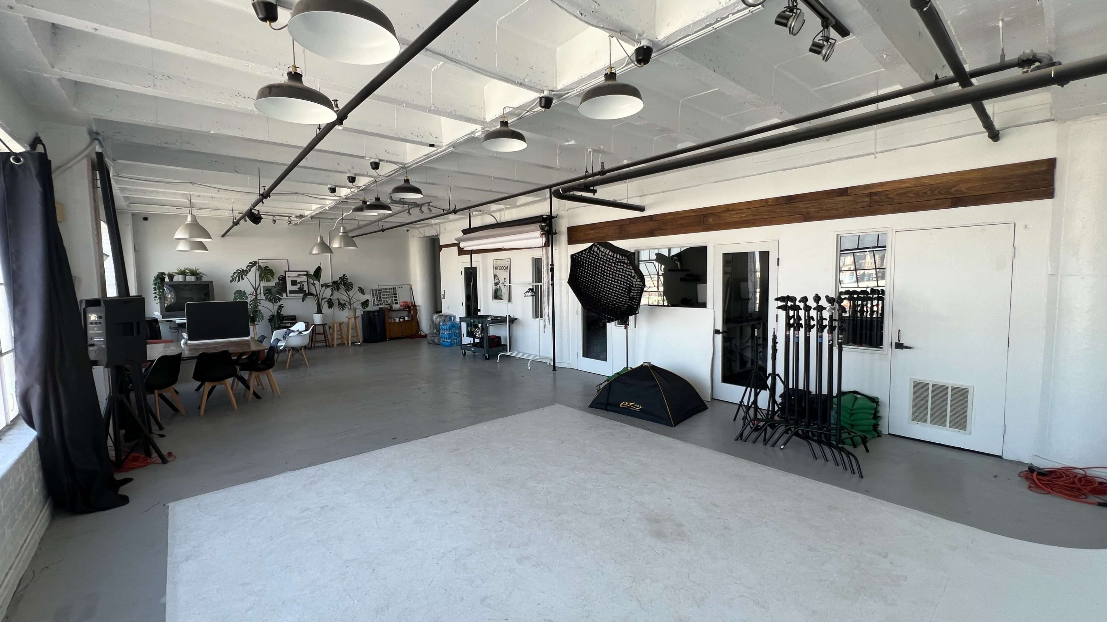 The image shows a spacious photography studio with a white floor, several large windows, a seating area, and various lighting equipment.