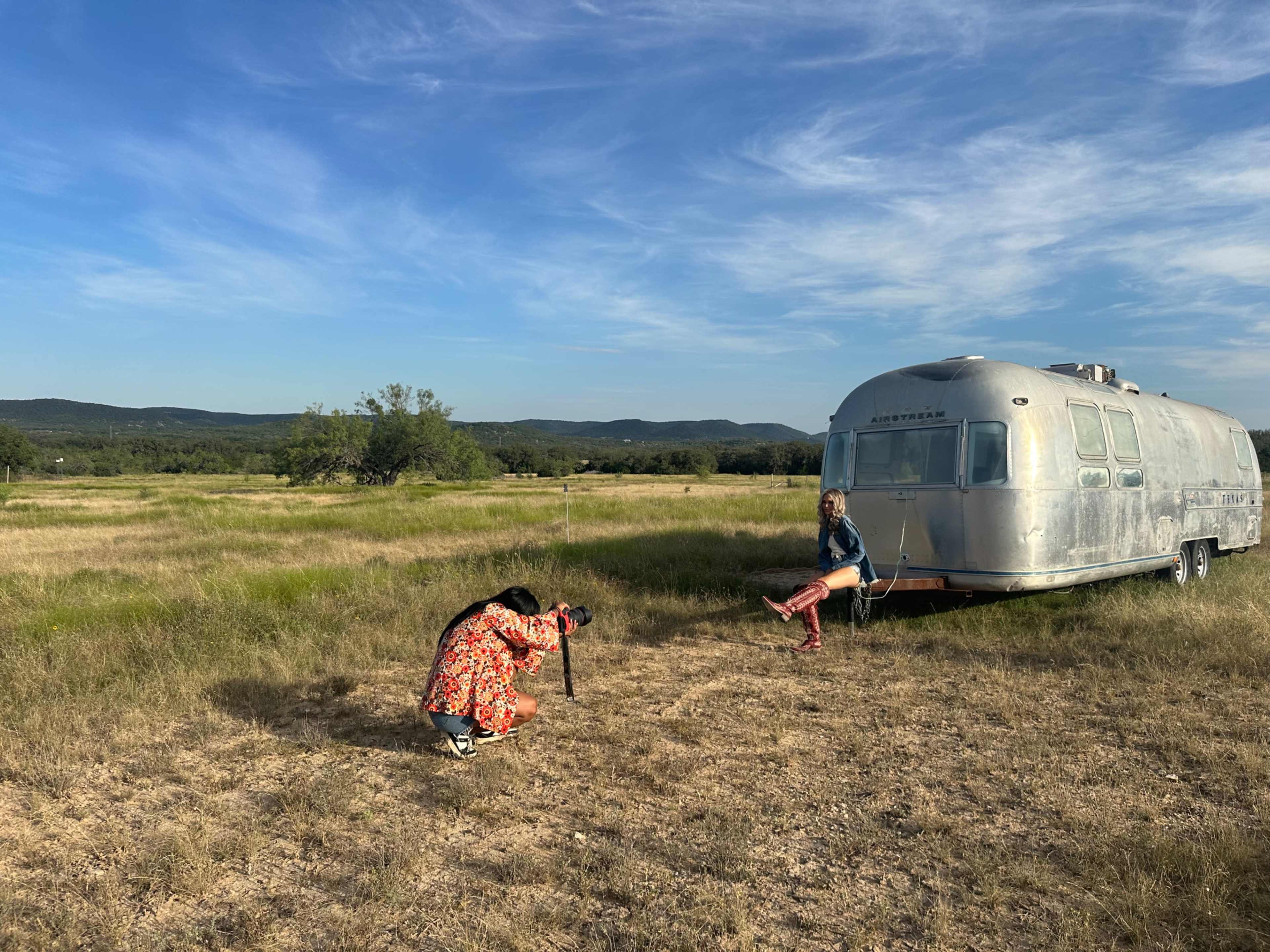 A person in a colorful outfit takes photos of another person sitting on the steps of a vintage trailer in a wide, open field.