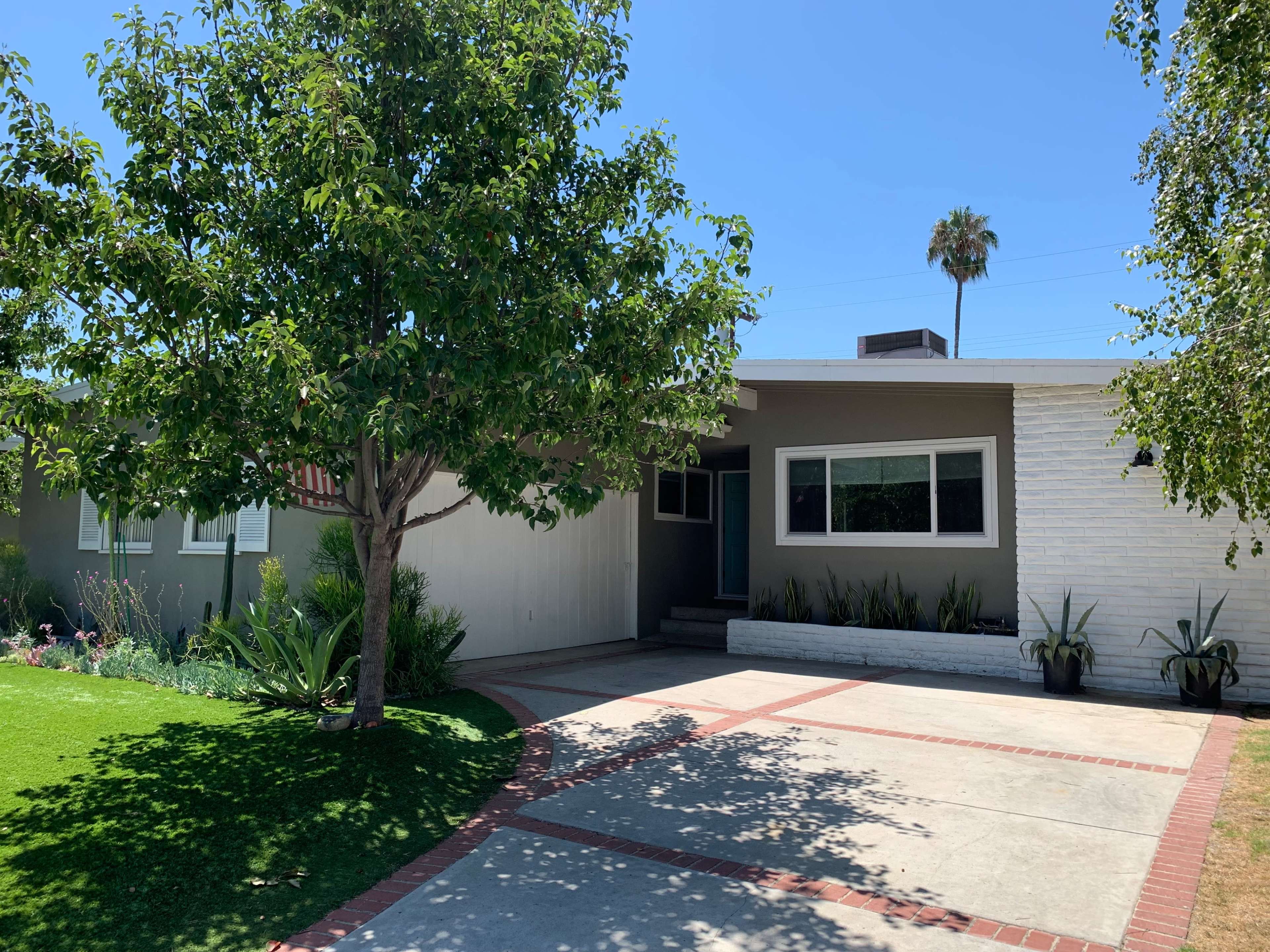 A single-story house features a concrete driveway, a front lawn with landscaping, and a large tree in the foreground.