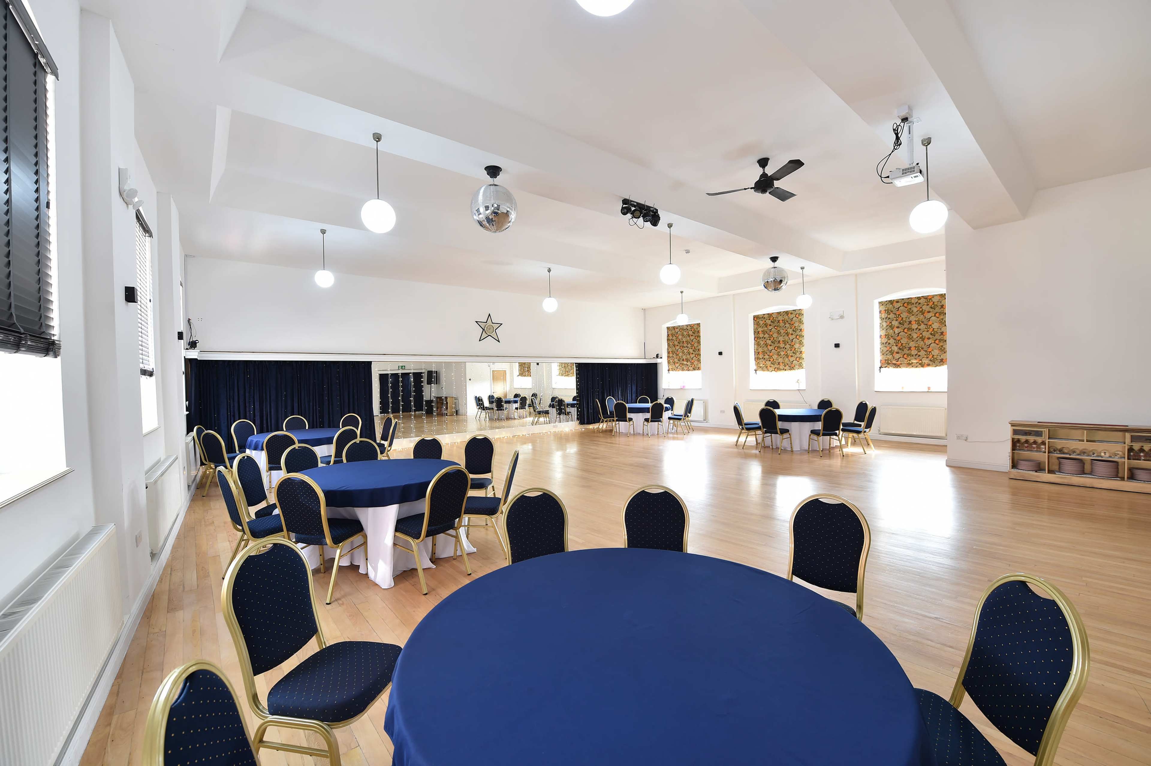 The image shows a spacious hall with several round tables set with blue tablecloths, surrounded by chairs, under bright overhead lighting.